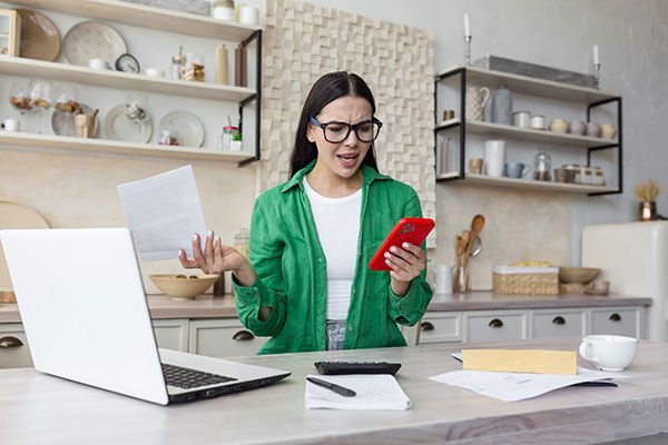 A woman is sitting at a table with a laptop and a cell phone.