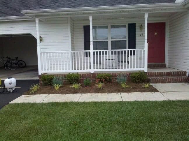 Front porch of a white house with red door, railing, steps, and small front garden beside a lawn