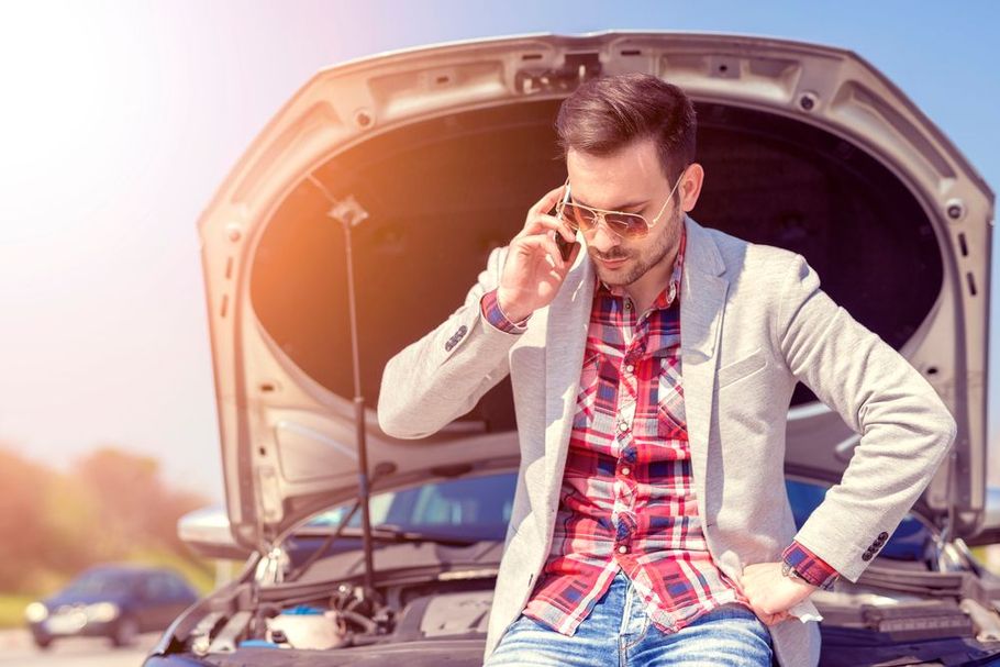 Man sitting on a car bumper with hood open, adjusting glasses and looking concerned.