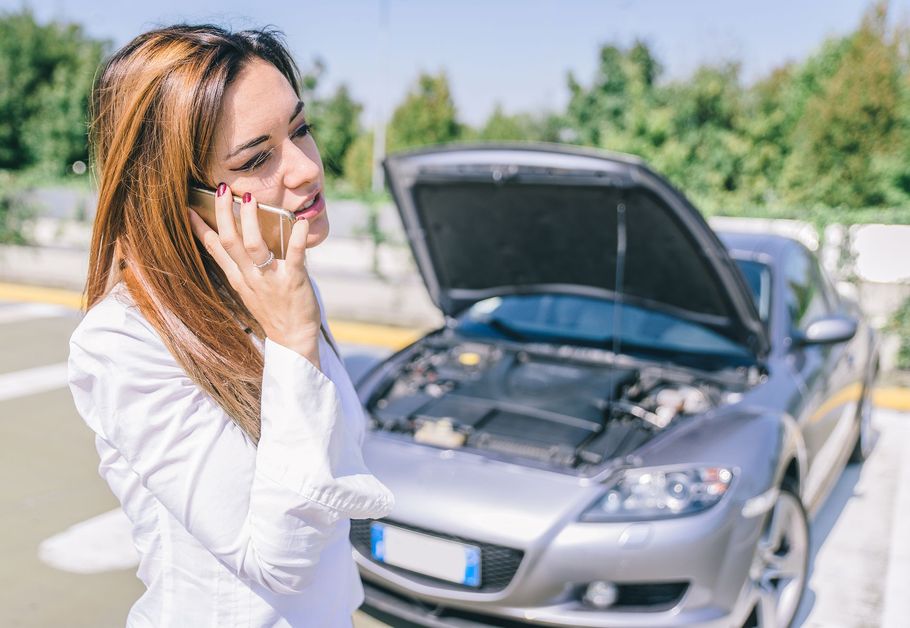 Woman on a phone beside a silver car with its hood open on a roadside.