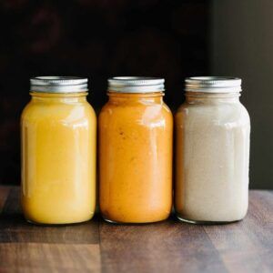 Three glass jars of creamy yellow, orange, and white sauce on a wooden surface.