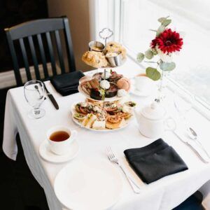 Tea set on a white table with desserts, black napkins, and a red flower centerpiece by a bright window