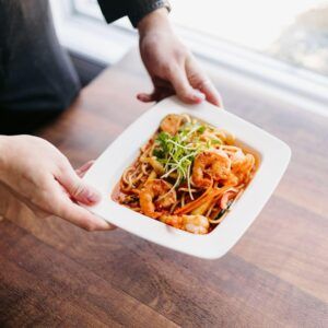Hands holding a white takeout bowl of pasta with shrimp and greens over a wooden table.