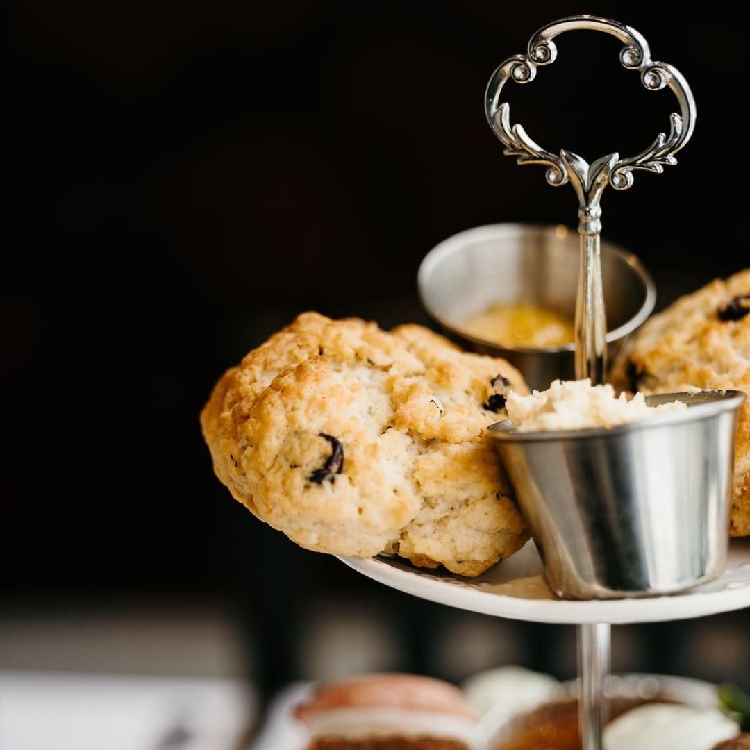 A tiered serving stand with scones and small metal cups of jam and clotted cream, set against a dark background.