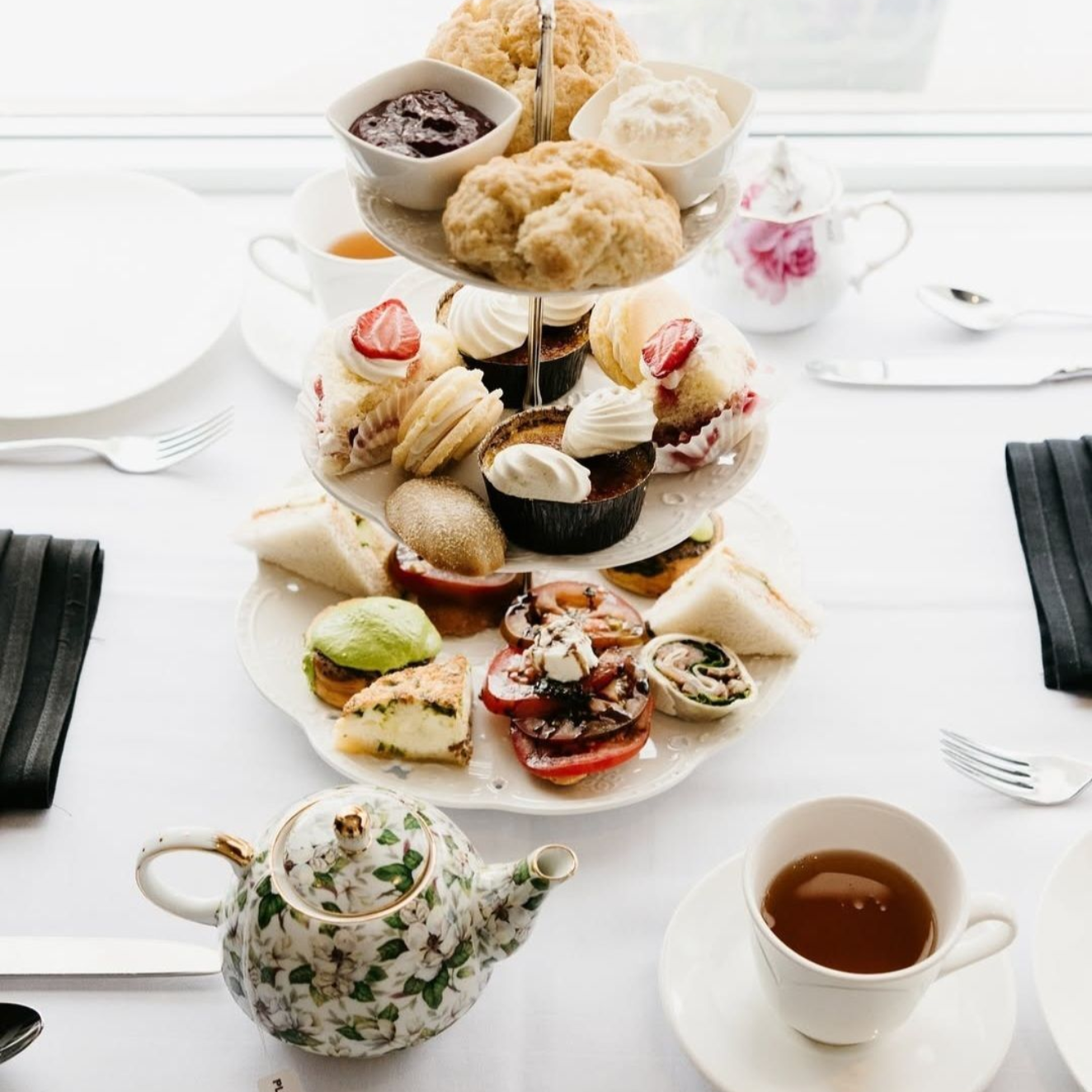 A tiered afternoon tea set with scones, pastries, and sandwiches on a white table with a floral teapot and teacups.