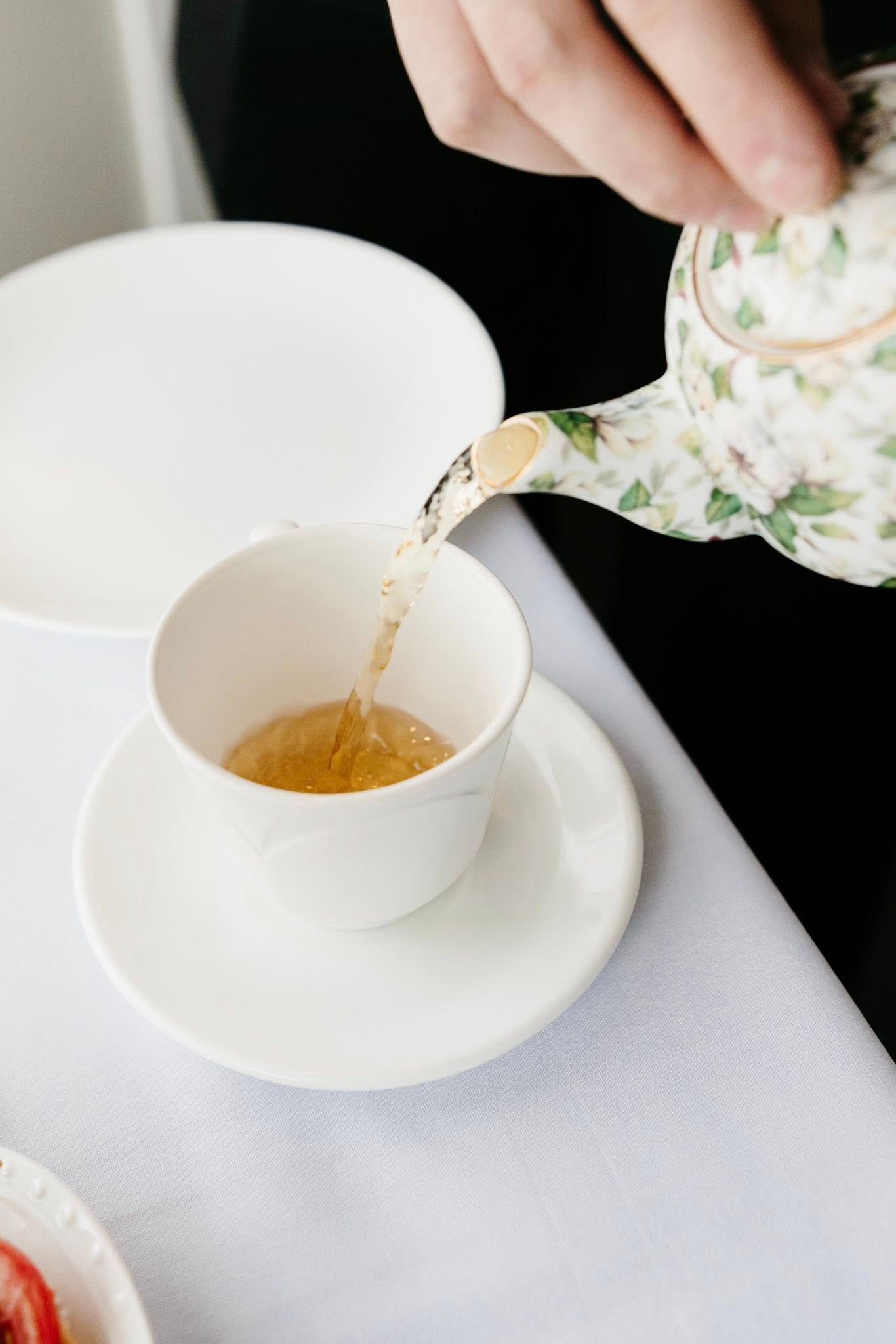 A floral teapot pours tea into a white teacup on a saucer atop a white tablecloth.
