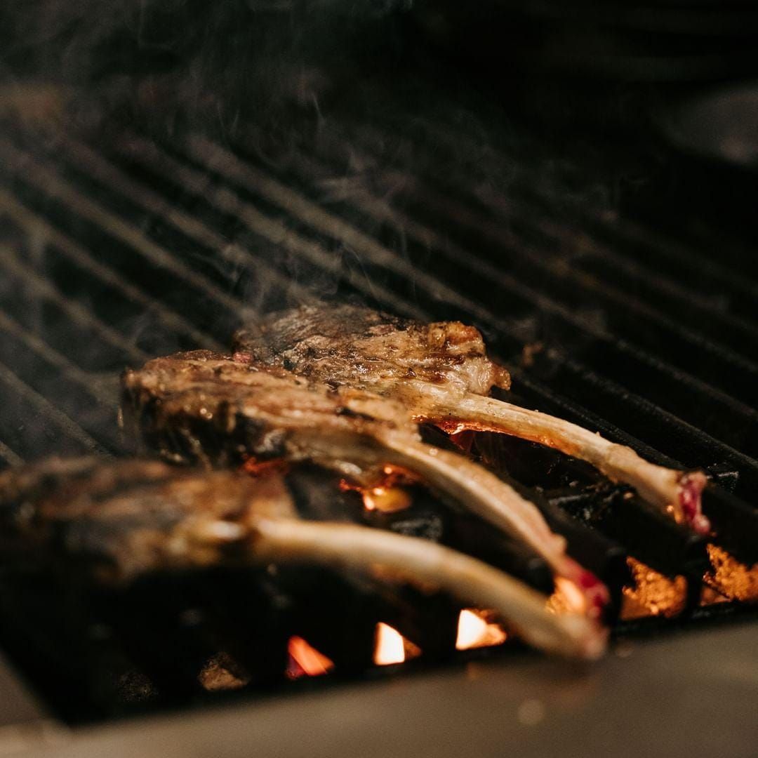 Lamb chops grilling over open flames on a barbecue, with charred meat and glowing embers.