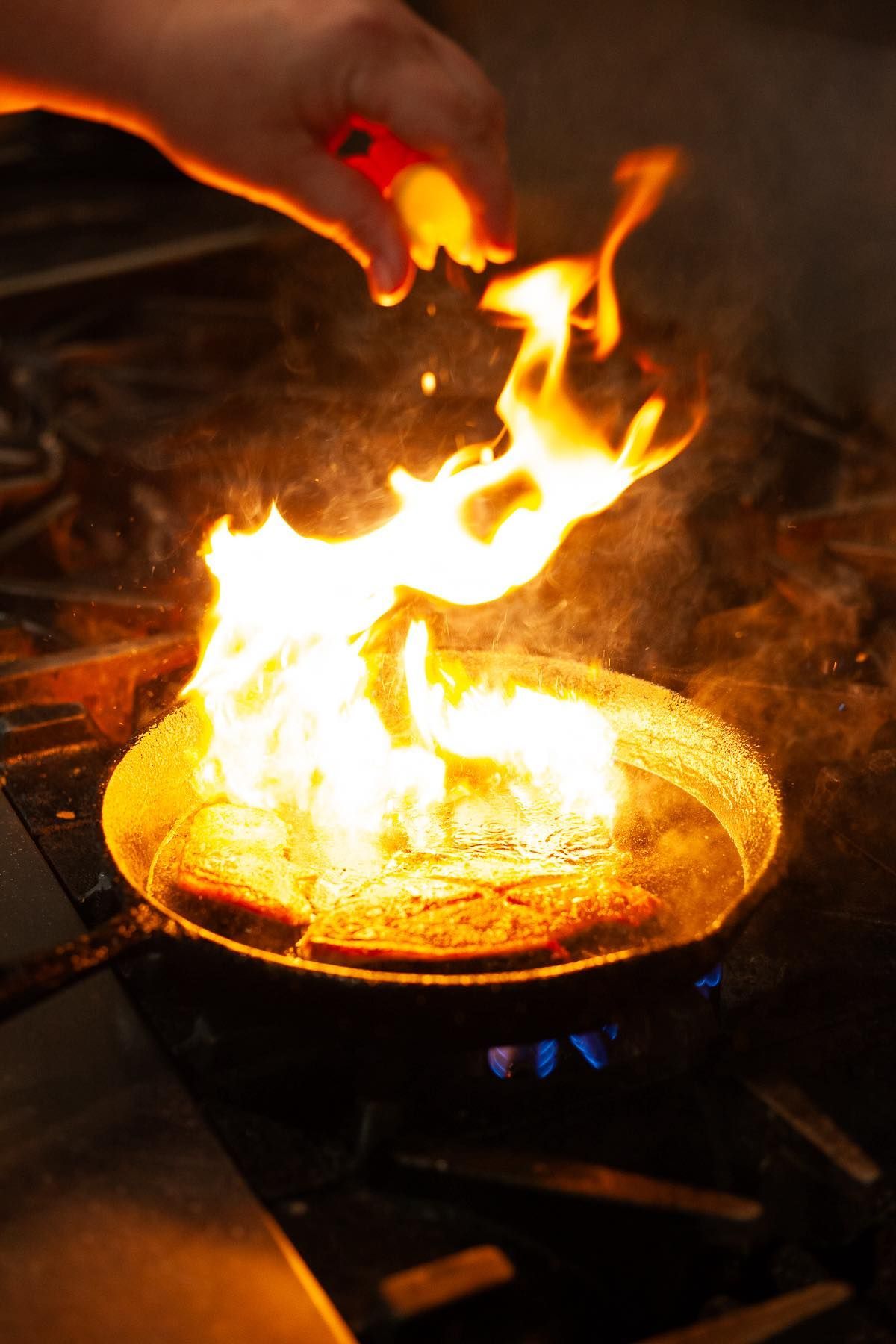 Hand adding seasoning to a flaming pan of food on a stovetop