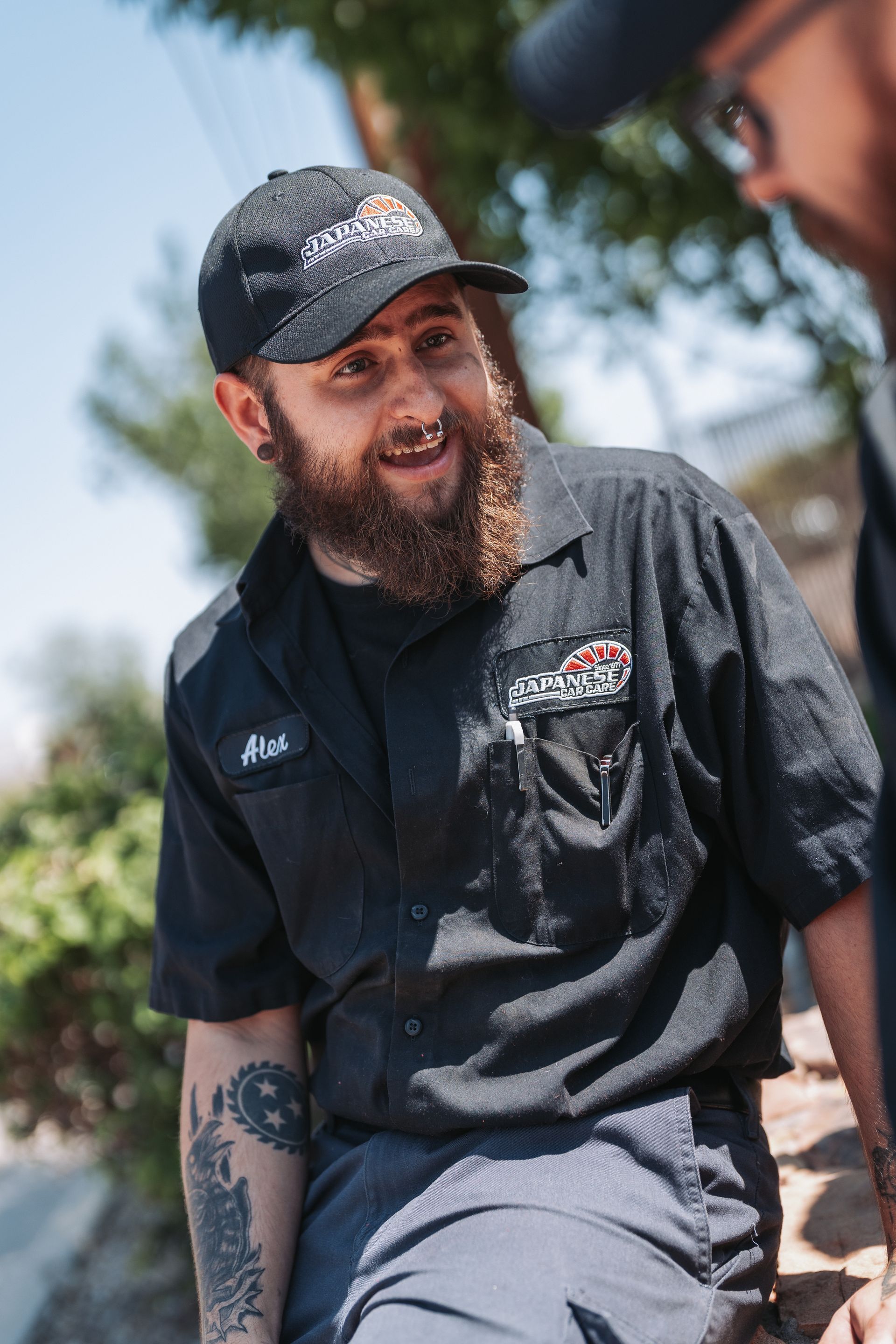 A man with a beard wearing a baseball cap and a black shirt is sitting next to another man. | Japanese Car Care
