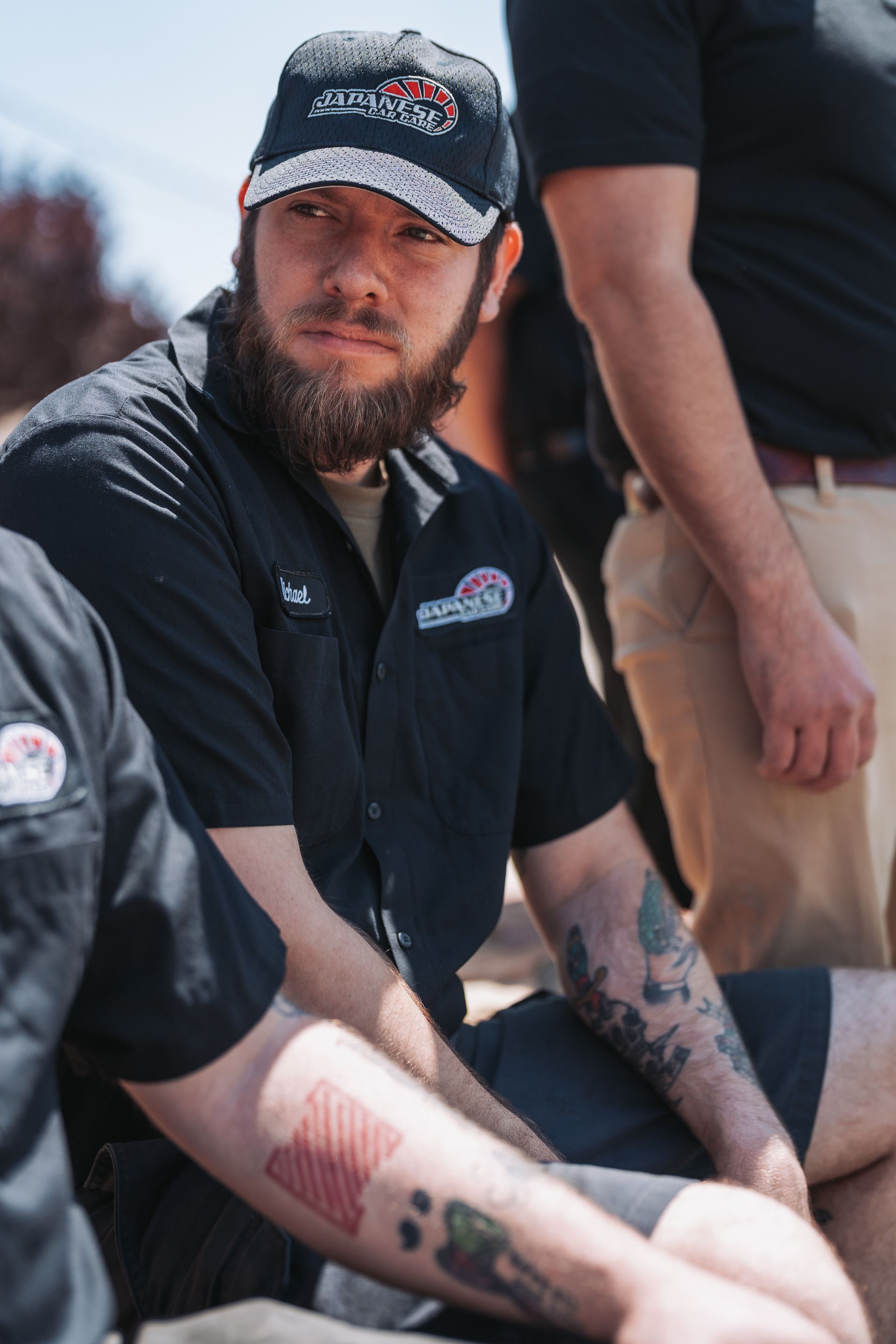 A man with a beard and tattoos is sitting on the ground wearing a baseball cap. | Japanese Car Care