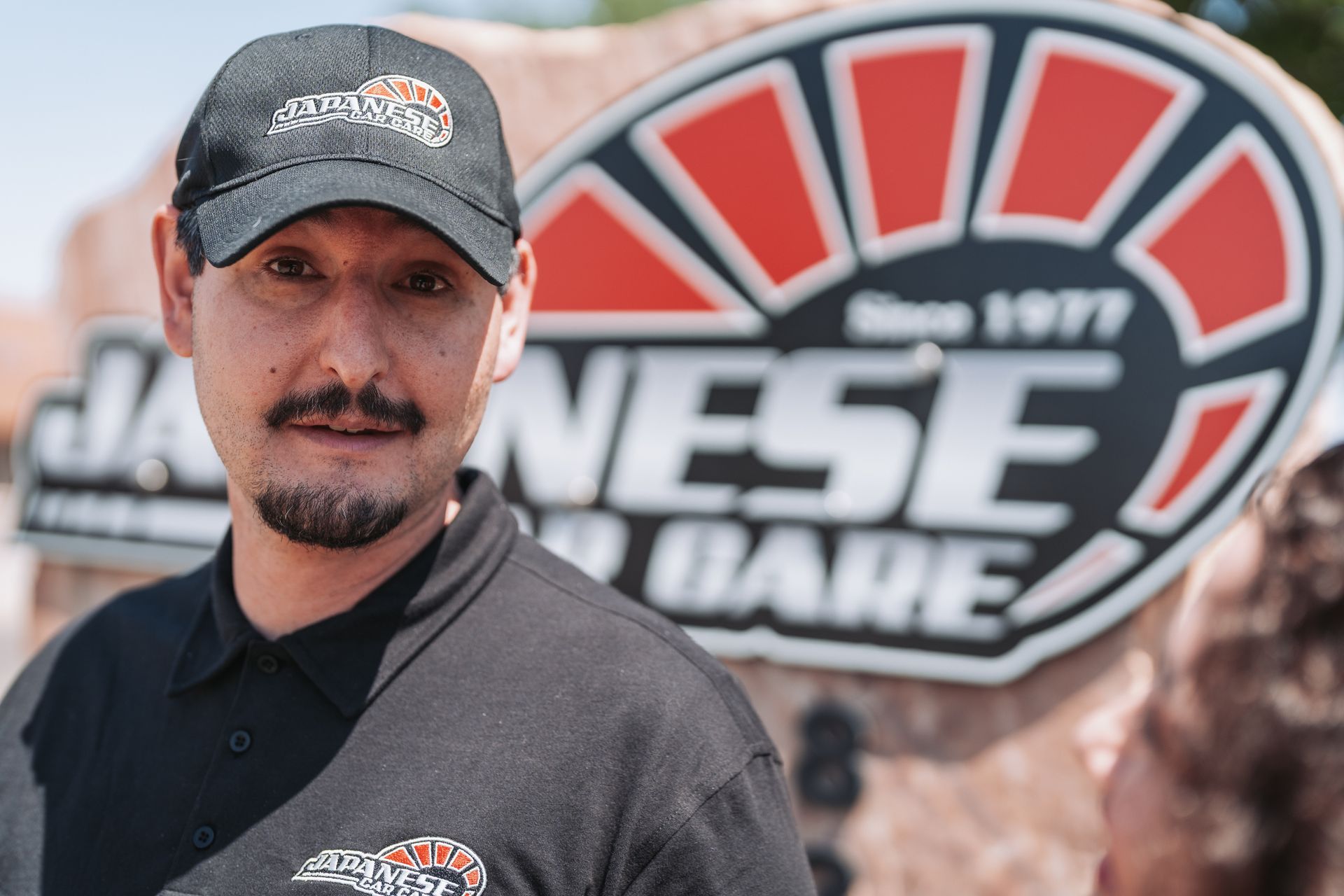 A man wearing a hat and a polo shirt is standing in front of a sign that says ' nes ' on it. | Japanese Car Care