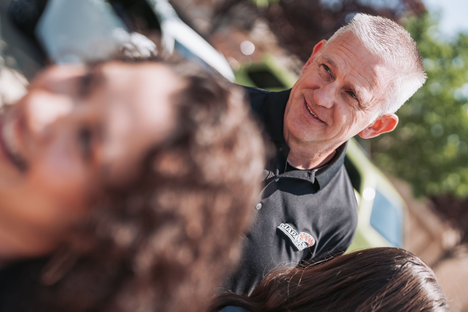 A man in a black shirt is talking to two women in front of a car. | Japanese Car Care