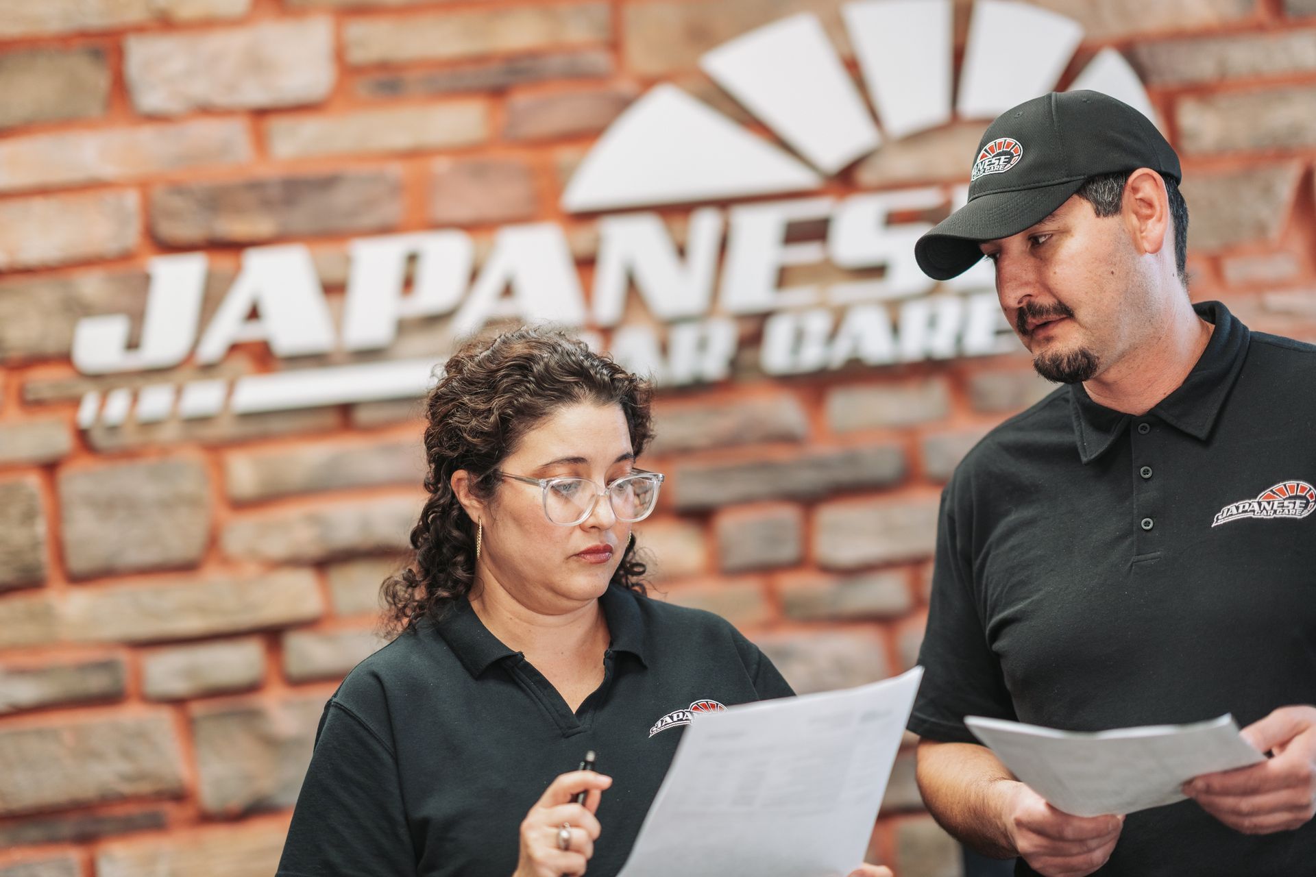 A man and a woman are looking at papers in front of a brick wall. | Japanese Car Care