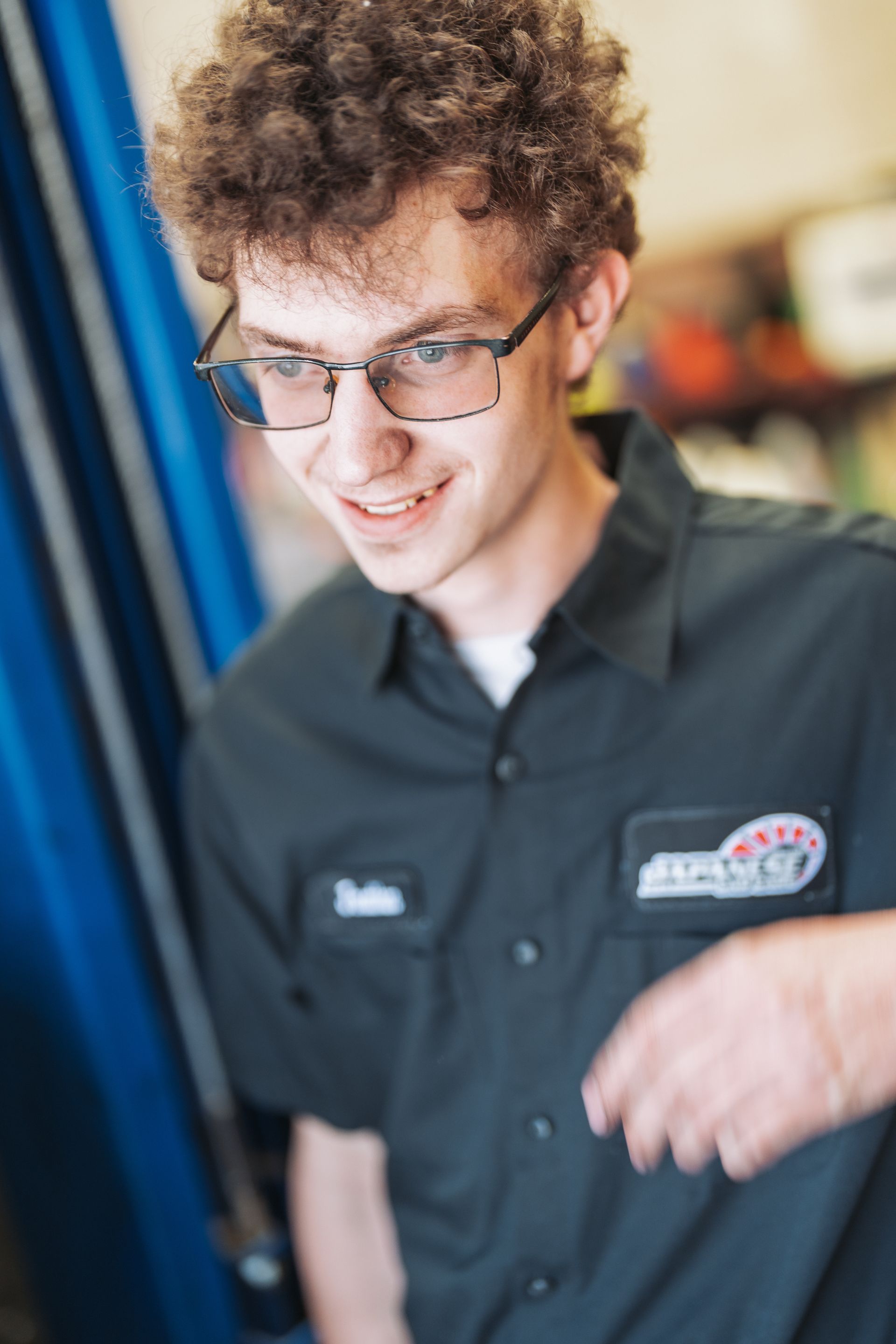 A young man wearing glasses and a black shirt is smiling in a garage. | Japanese Car Care