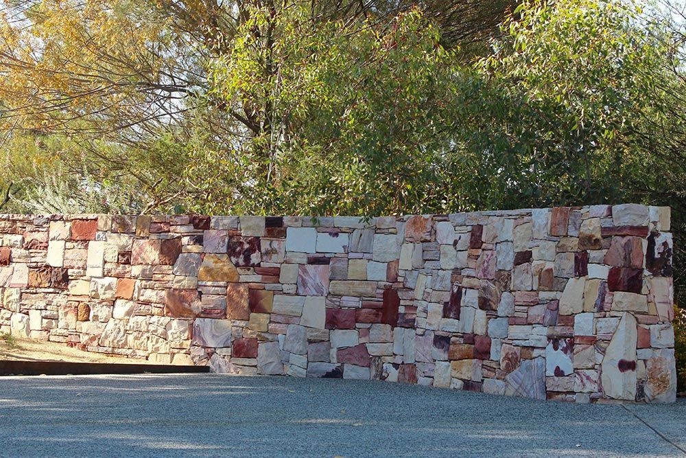 Mosaic stone wall along a paved path, with trees and greenery behind it in daylight