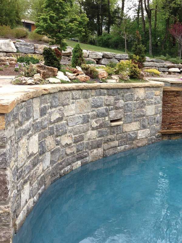 Curved stone retaining wall beside a blue pool with landscaped rocks and greenery