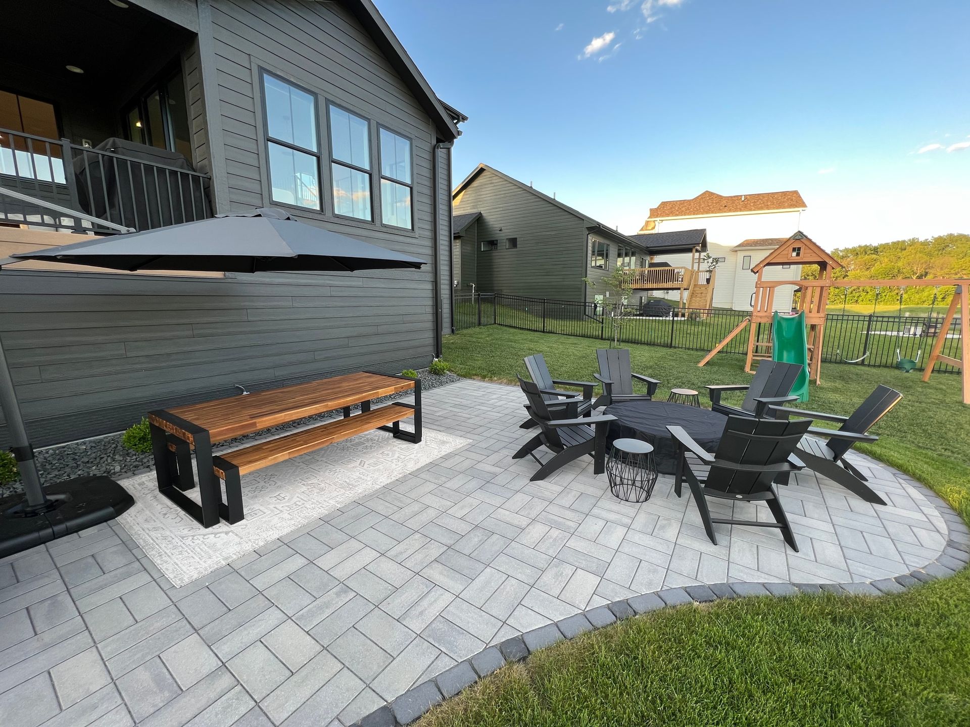 A modern gray stone patio with a wooden dining table, a black fire pit surrounded by outdoor chairs, and a yard.