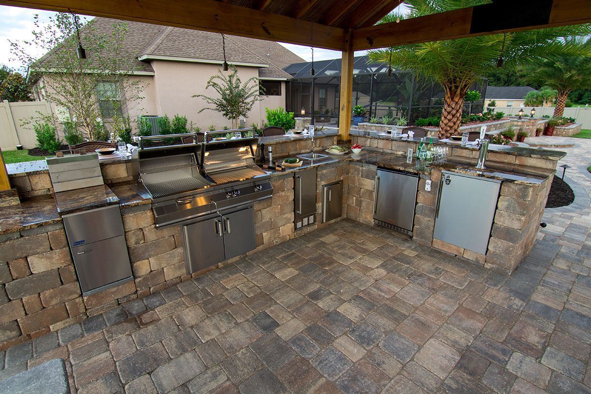 Outdoor stone kitchen under a wooden pergola with grill, sink, cabinets, and potted plants
