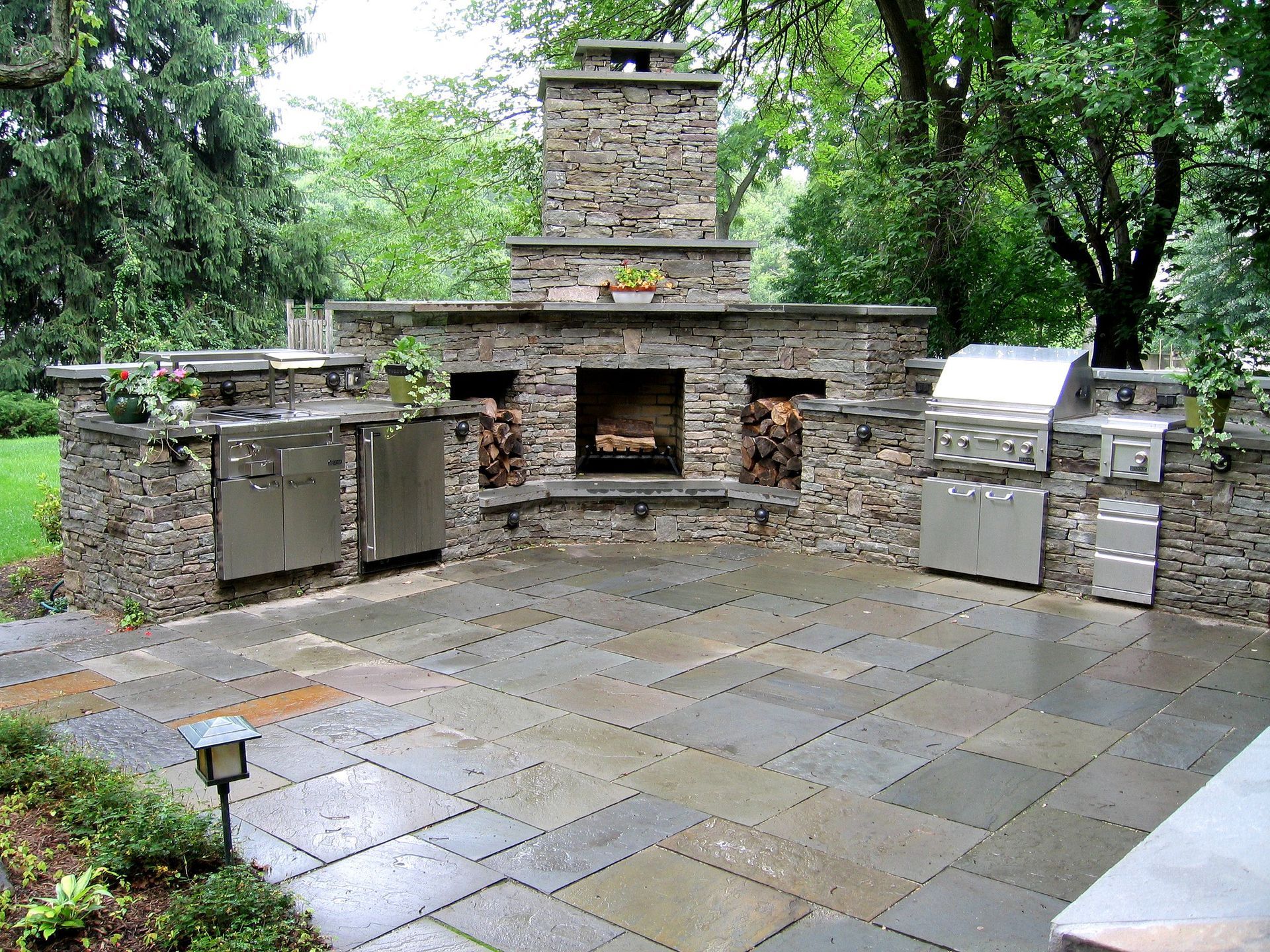 Stone outdoor fireplace and grill area with a paved patio surrounded by trees