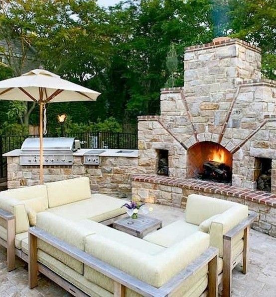 Outdoor patio with cream sectional seating, umbrella, grill, and stone fireplace with a lit fire.