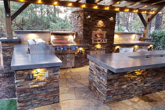 Covered outdoor stone kitchen with built-in grills, counters, and warm overhead lights at dusk