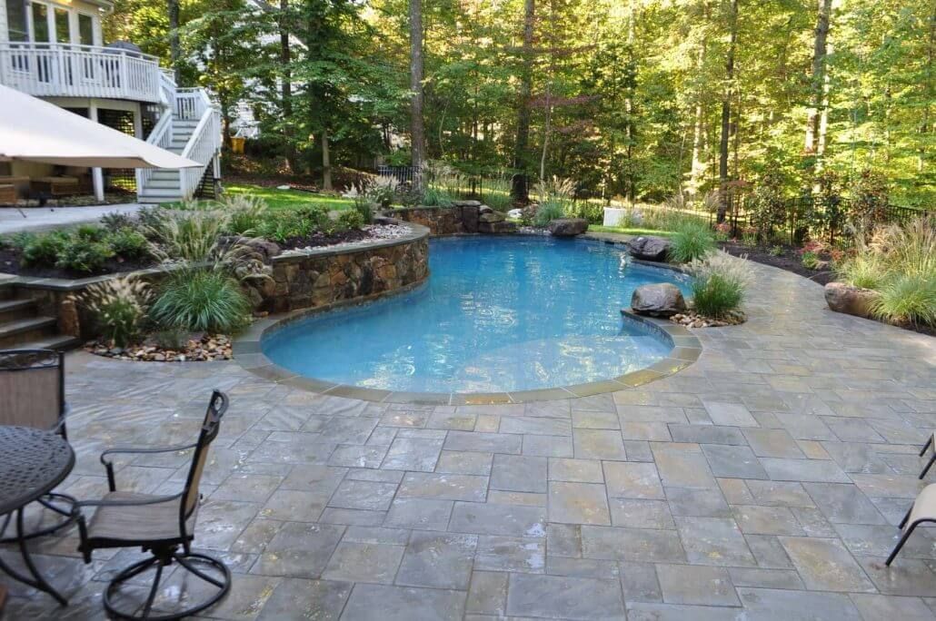 Backyard stone patio with a small blue swimming pool surrounded by trees and garden landscaping