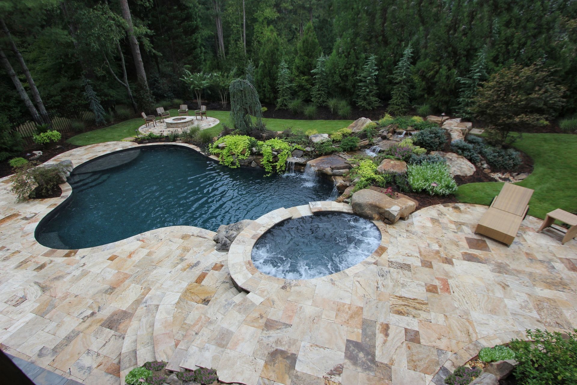 Backyard stone patio with a dark blue pool, bubbling spa, and lush landscaping