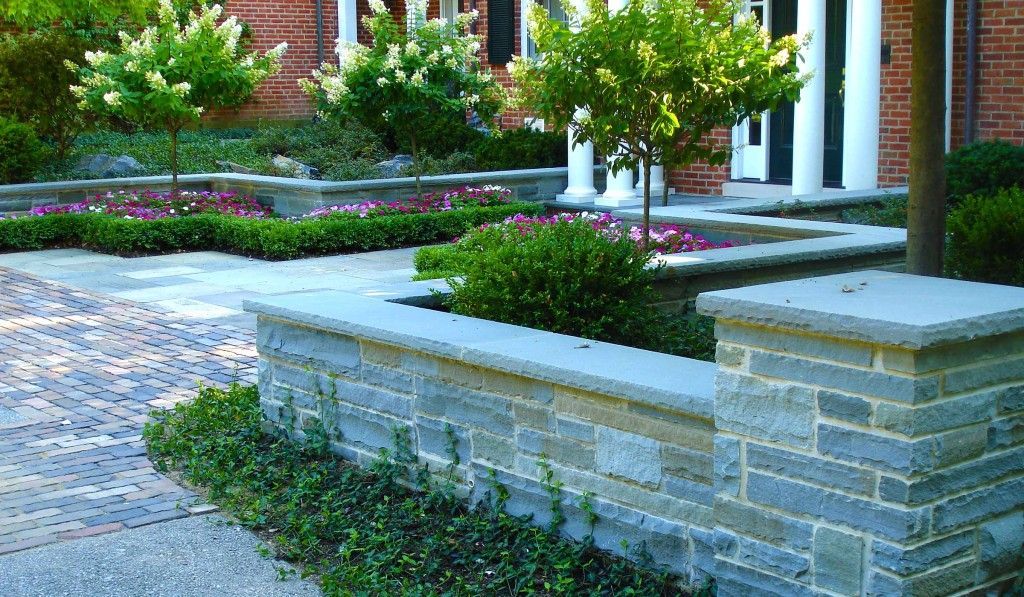 Landscaped brick patio with stone planters, shrubs, and a walkway beside a building entrance