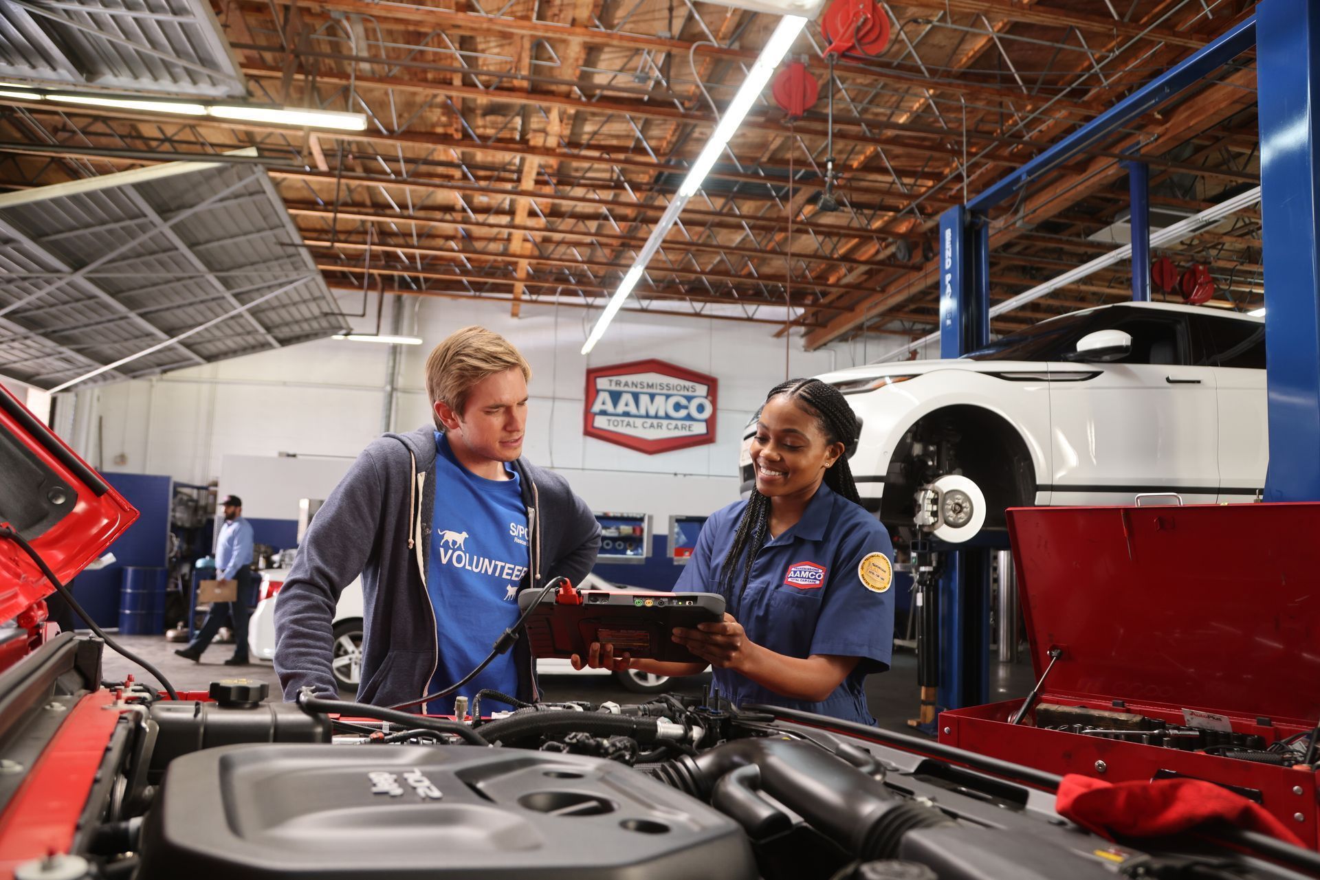 AAMCO mechanic showing a man a tablet while working on a car engine in an AAMCO garage.