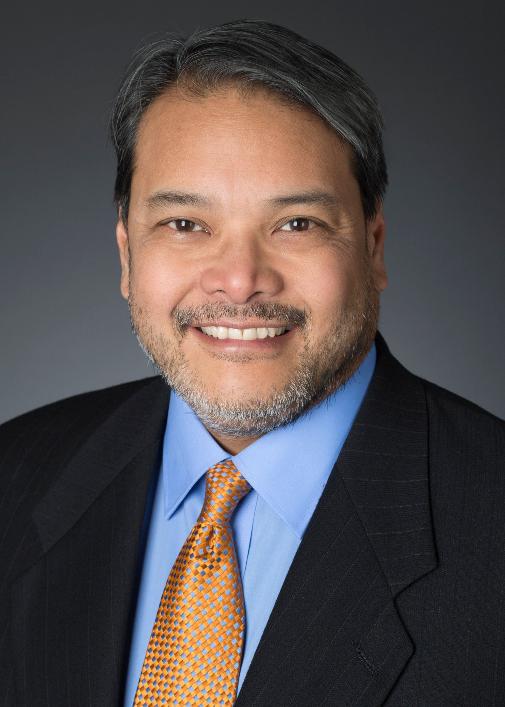 Man in blue shirt, orange tie, and black suit smiles at the camera, against a neutral backdrop.