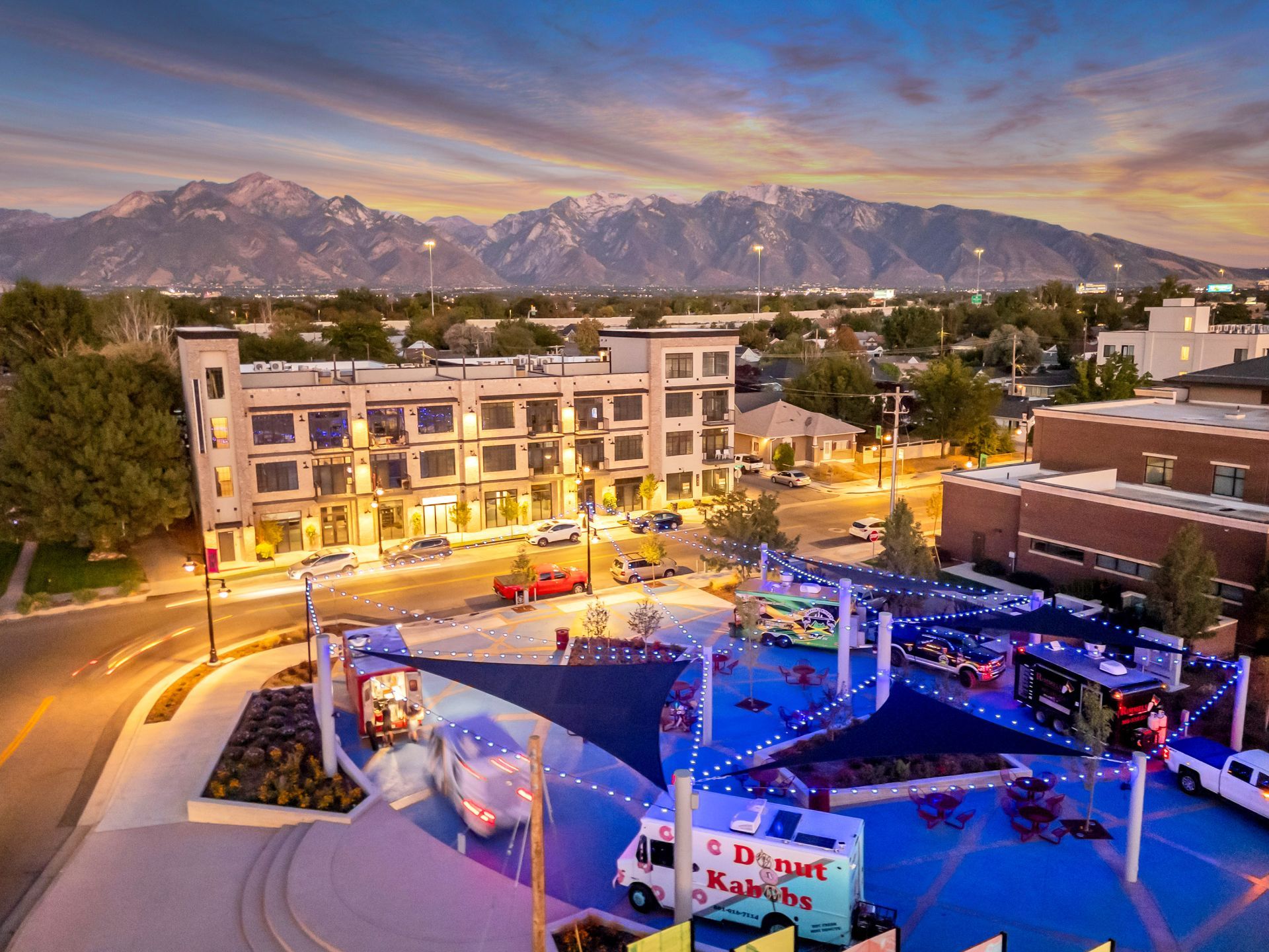 Food trucks gather in a plaza with shade sails. Apartments and mountains in the background.