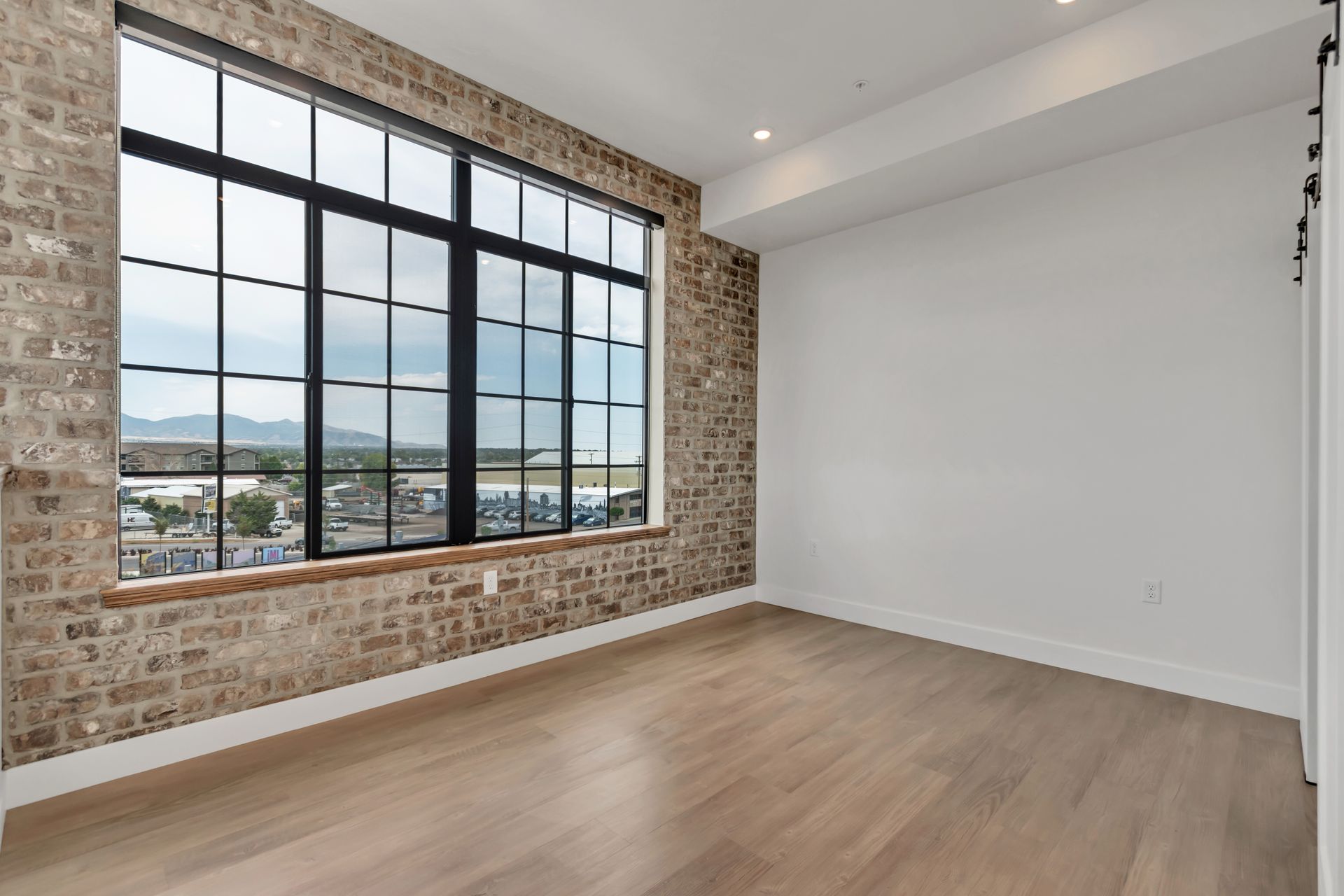 Empty room with brick wall, large window with a view, wood floor, and white walls.