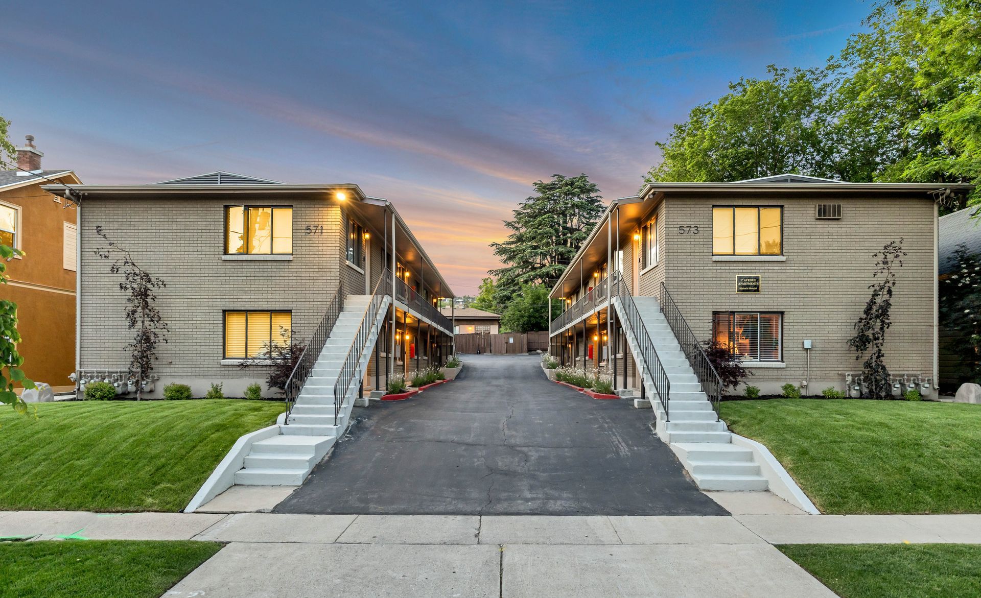 Two-story tan apartment buildings face each other, connected by a paved driveway. Lush green bushes and blue sky.
