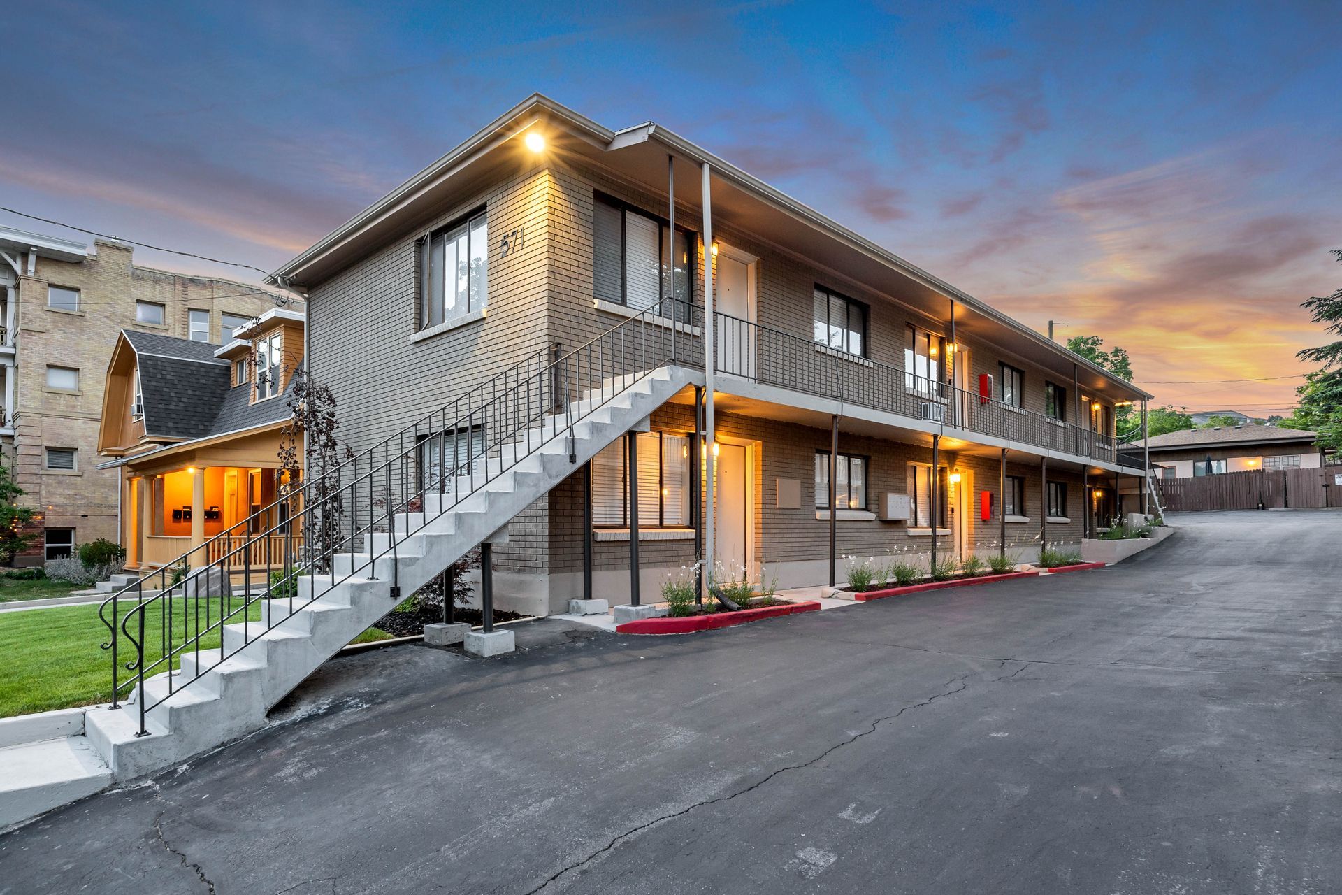 Two-story apartment building with exterior stairs and asphalt parking area under a blue sky.