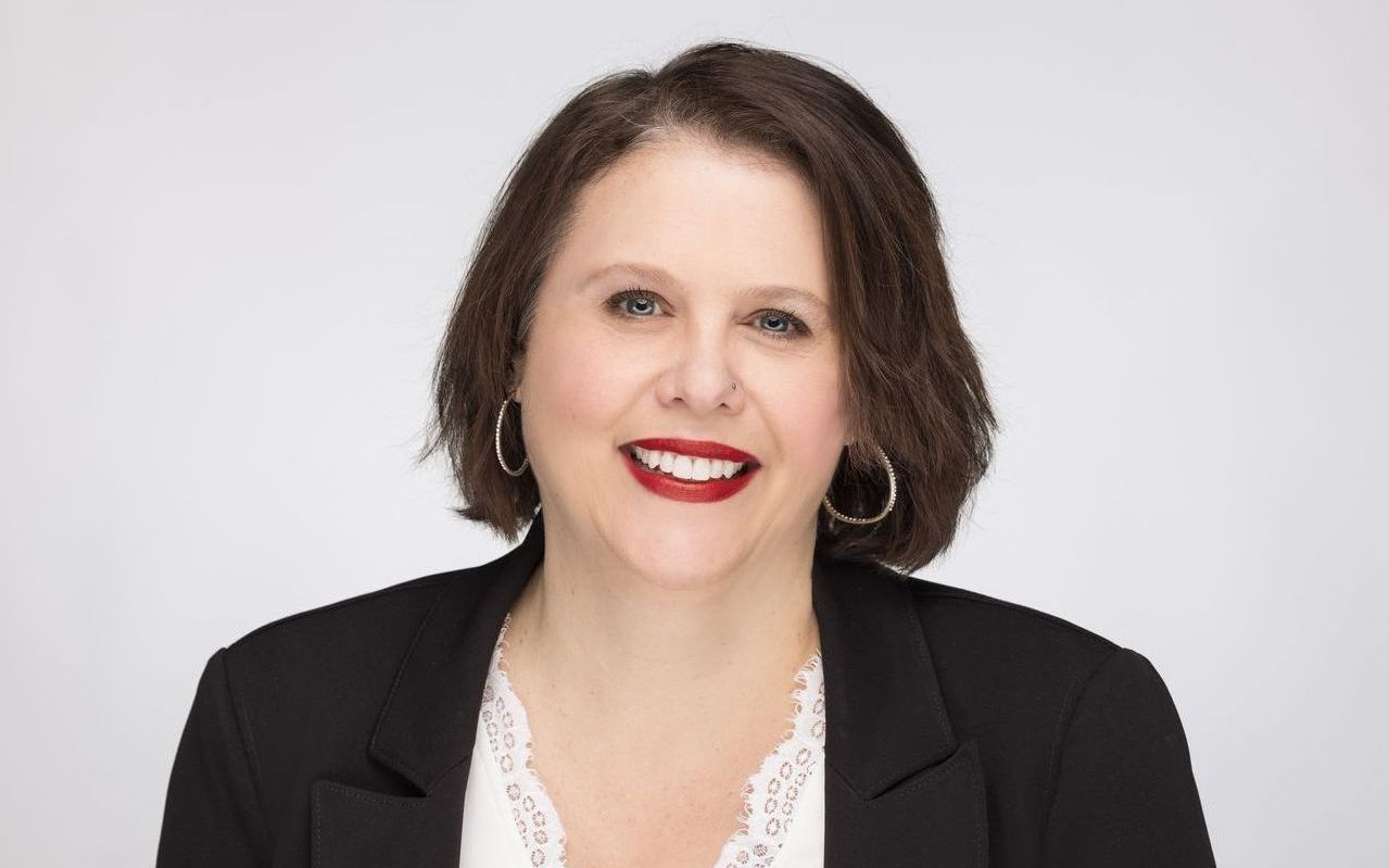 A woman wearing a black jacket and red lipstick is smiling for the camera.