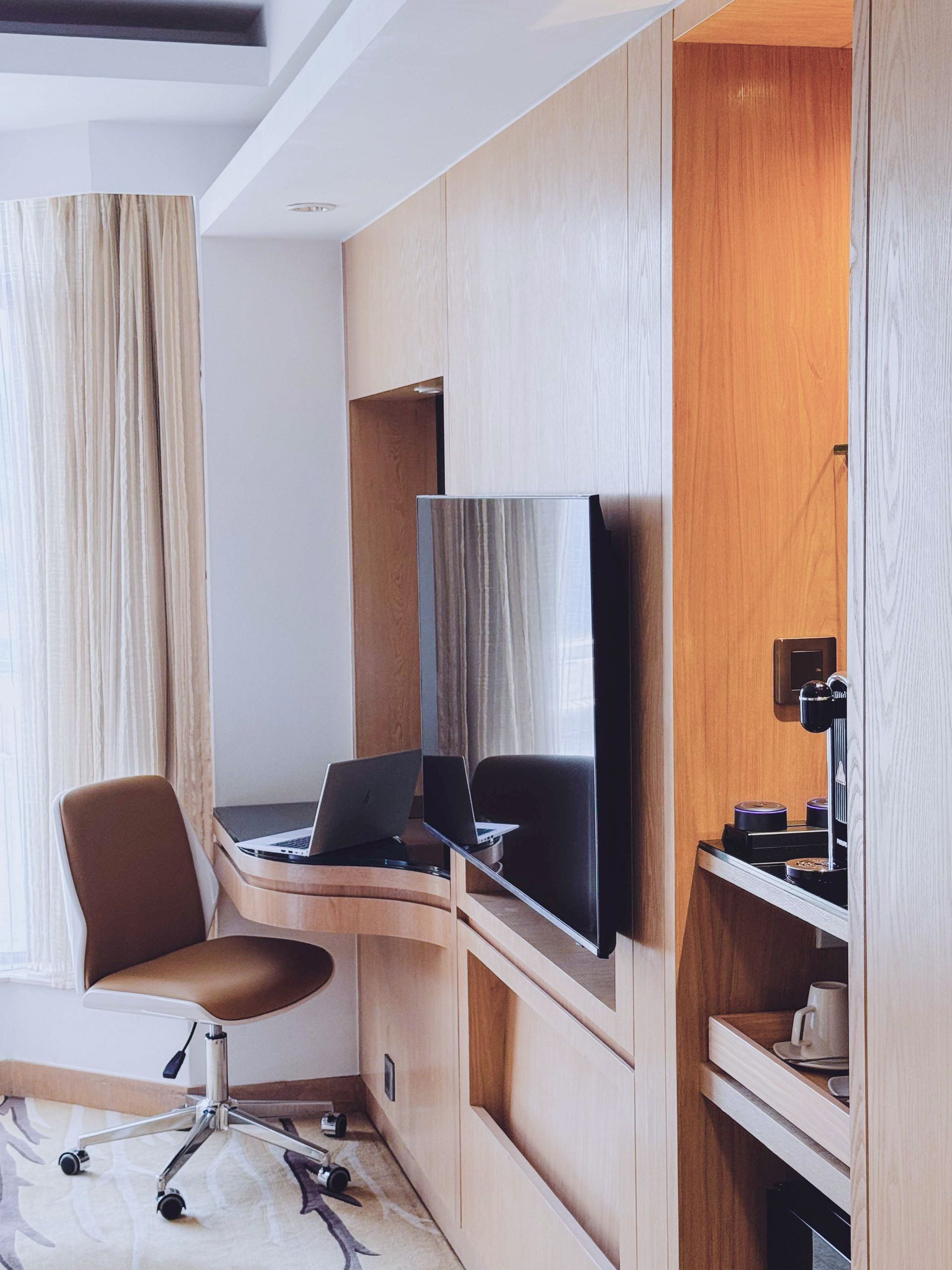 A modern hotel room with a wooden desk, a laptop, a swivel chair, a wall-mounted TV, and a built-in coffee station.