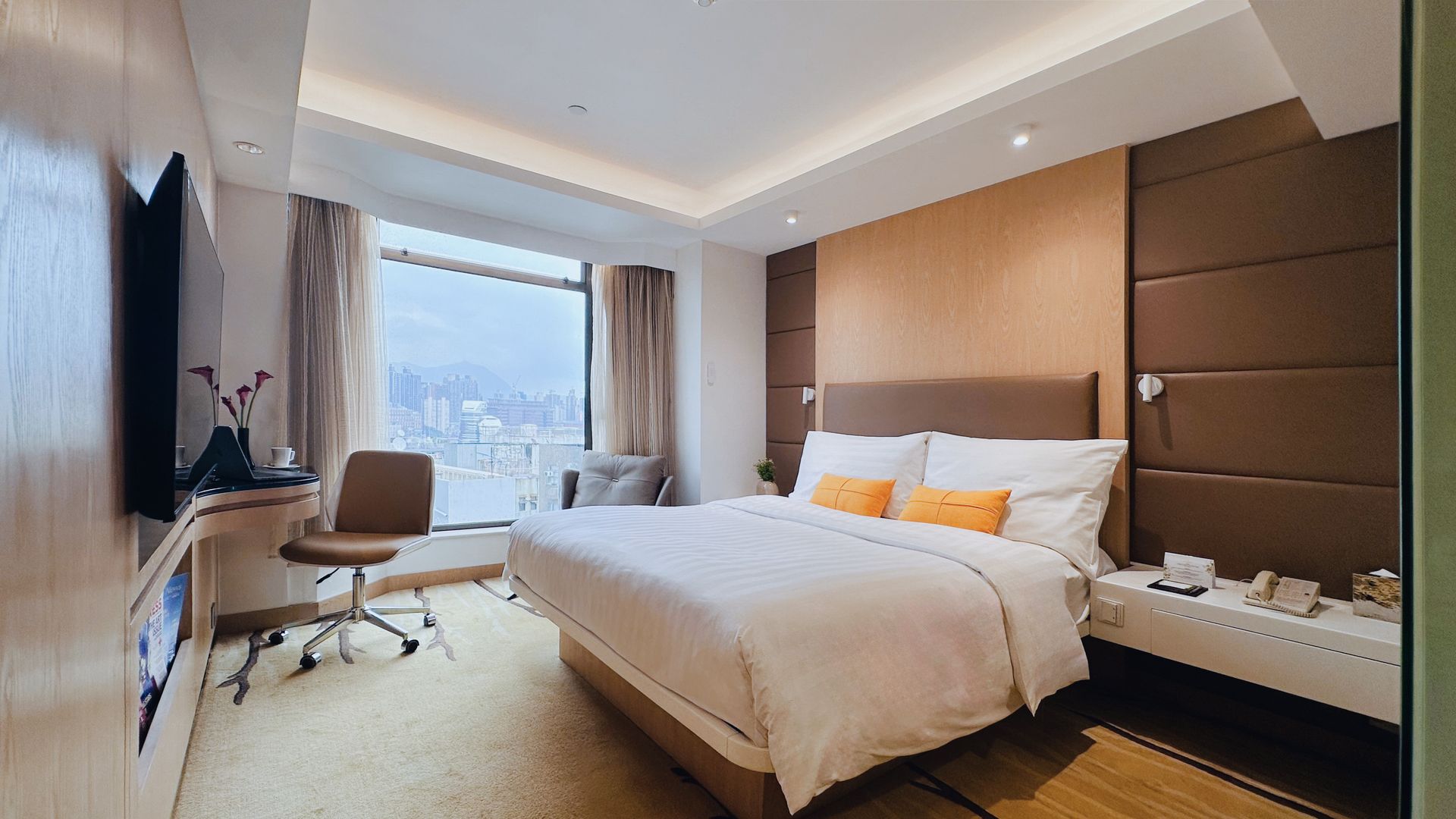 Hotel room with a bed, desk, and large window overlooking a city. Warm color scheme.
