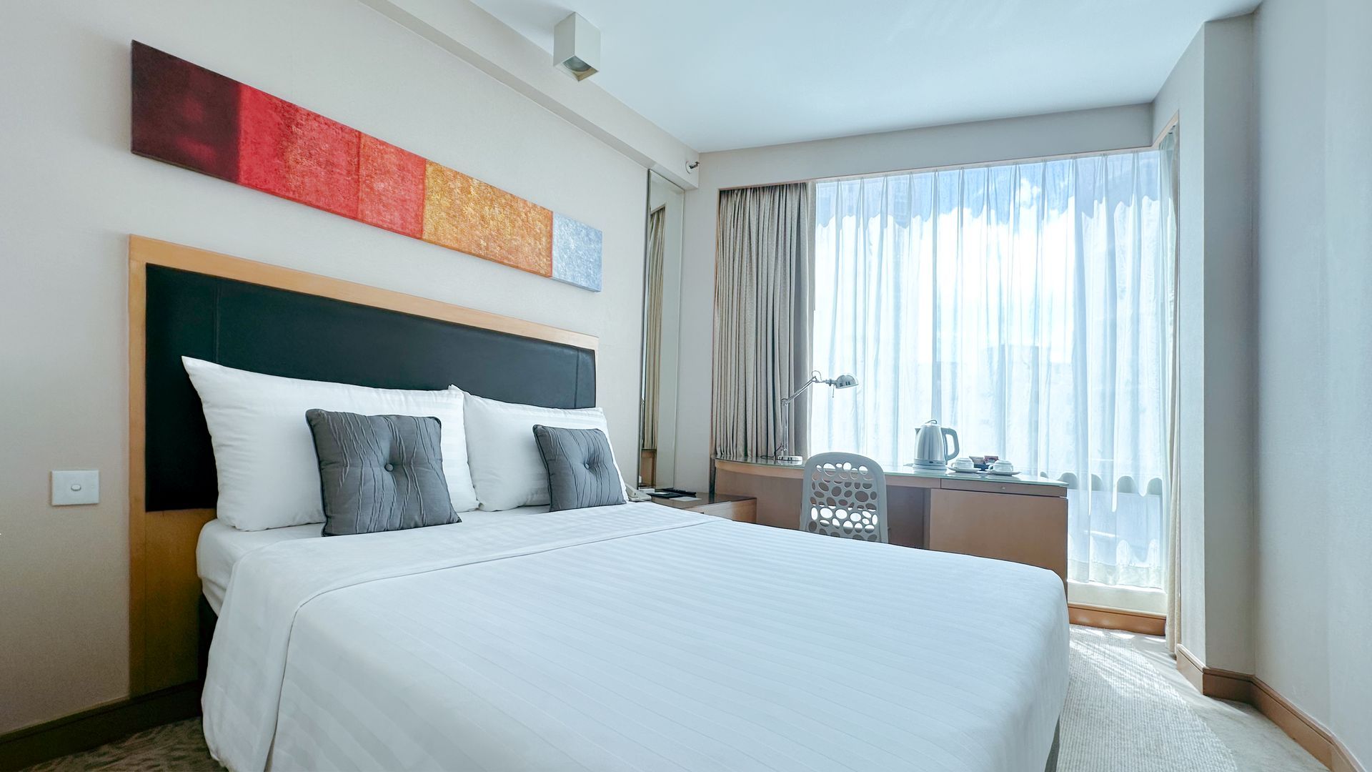 Hotel room with bed, desk with laptop, window with curtains, and art on the wall.
