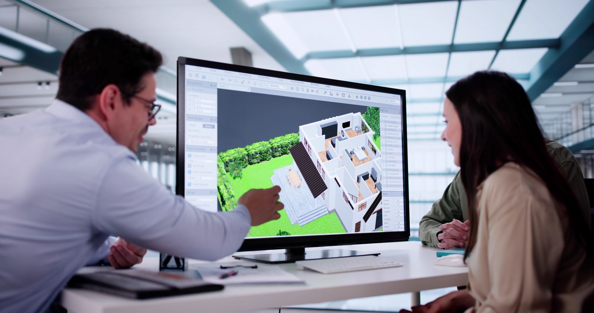 Deux personnes observent une maquette architecturale 3D d'une maison affichée sur un grand écran d'ordinateur dans un bureau.