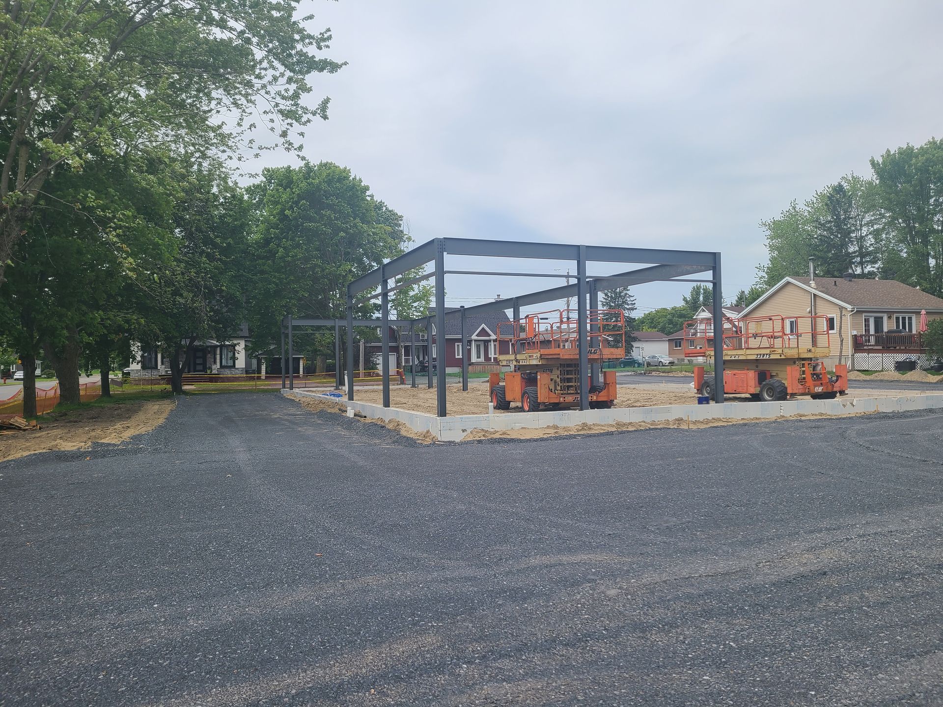 Structure métallique sur un terrain gravillonné, avec engins de chantier et maisons environnantes, dans un quartier résidentiel.