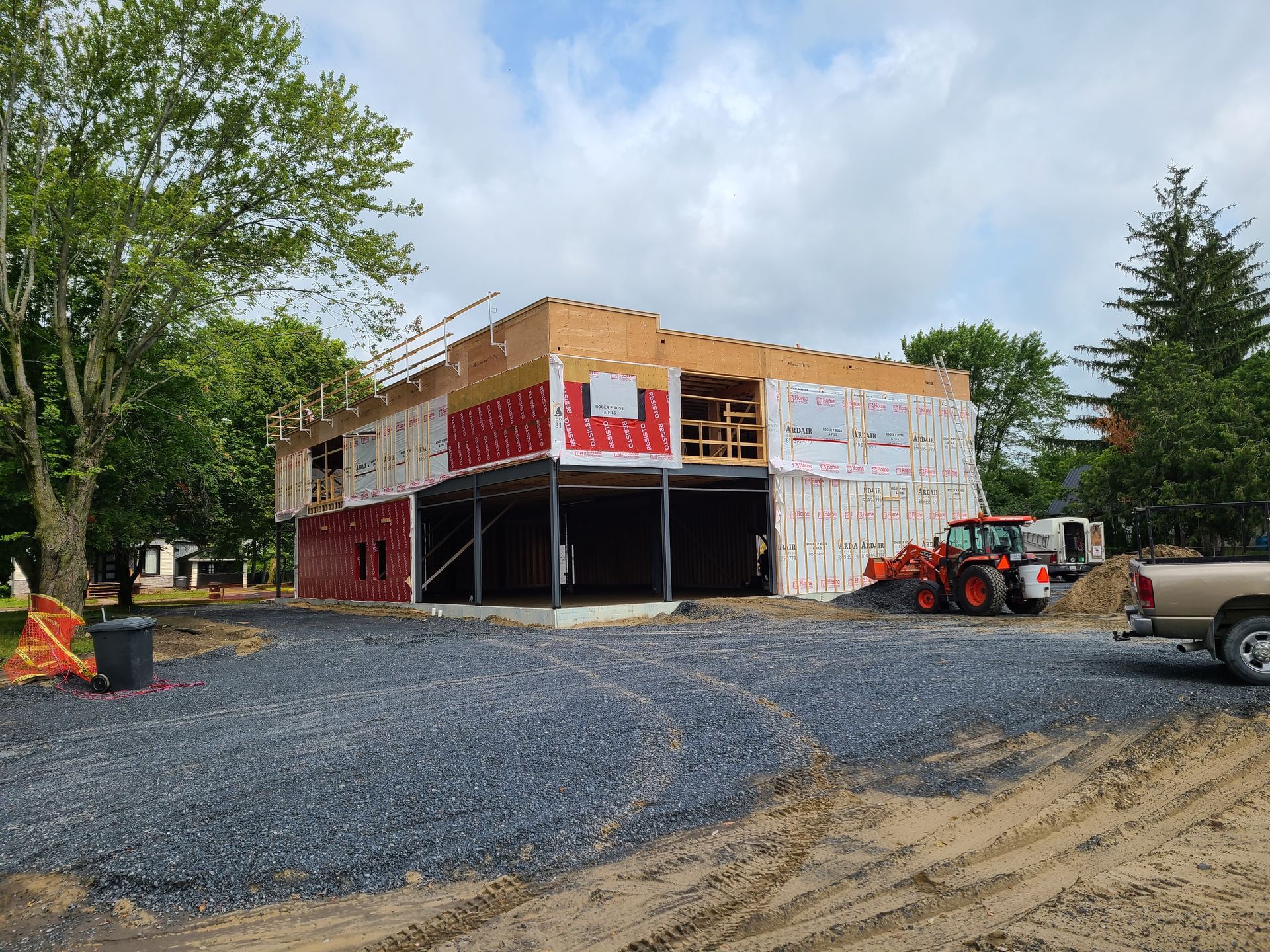 Une structure de bâtiment à deux étages partiellement construite, recouverte d'une bâche de protection, sur un chantier avec du gravier et un tracteur.