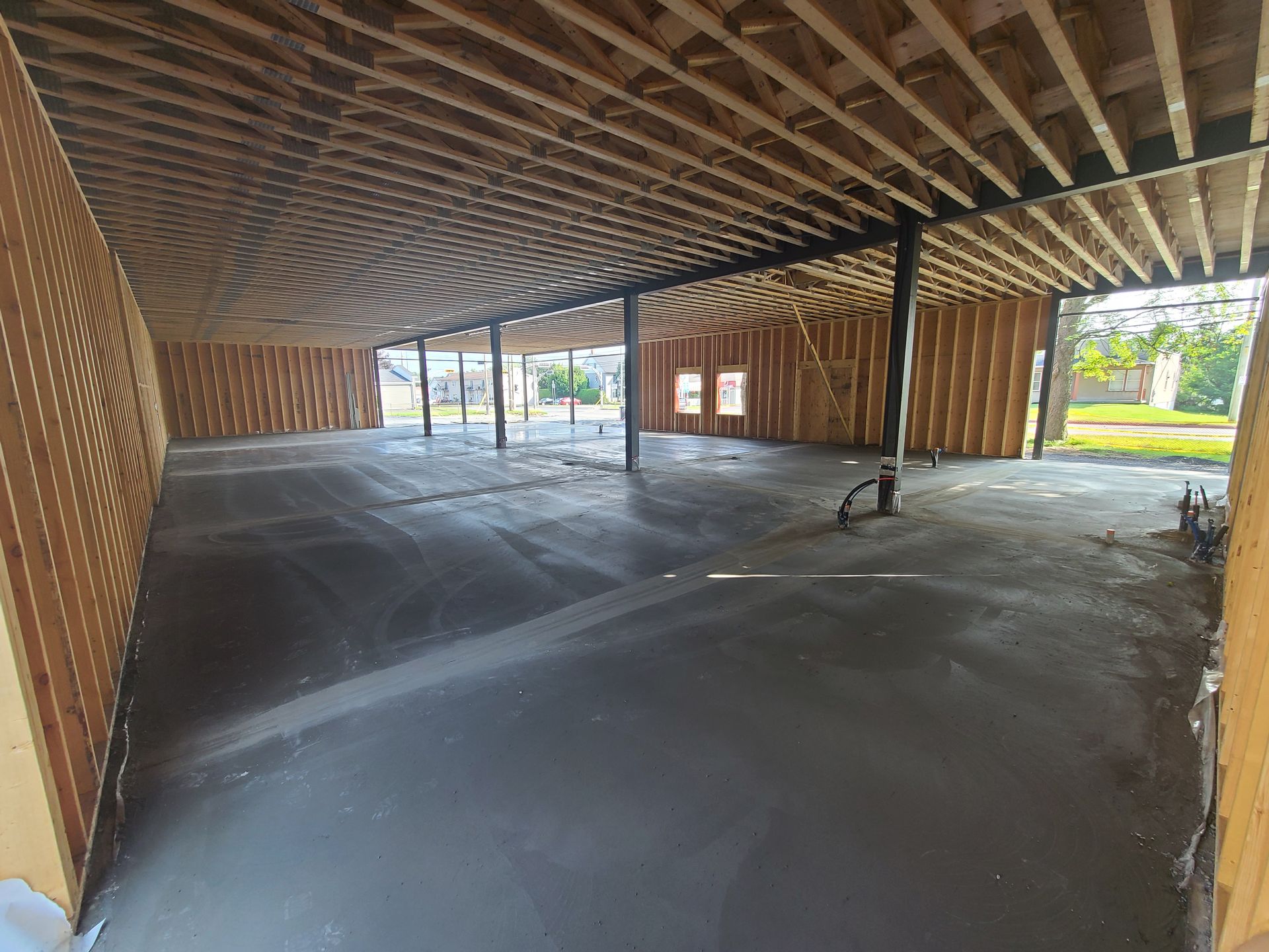 Vue intérieure d'un bâtiment en construction, montrant les montants muraux et les solives de plafond en bois apparents au-dessus d'un sol en béton.