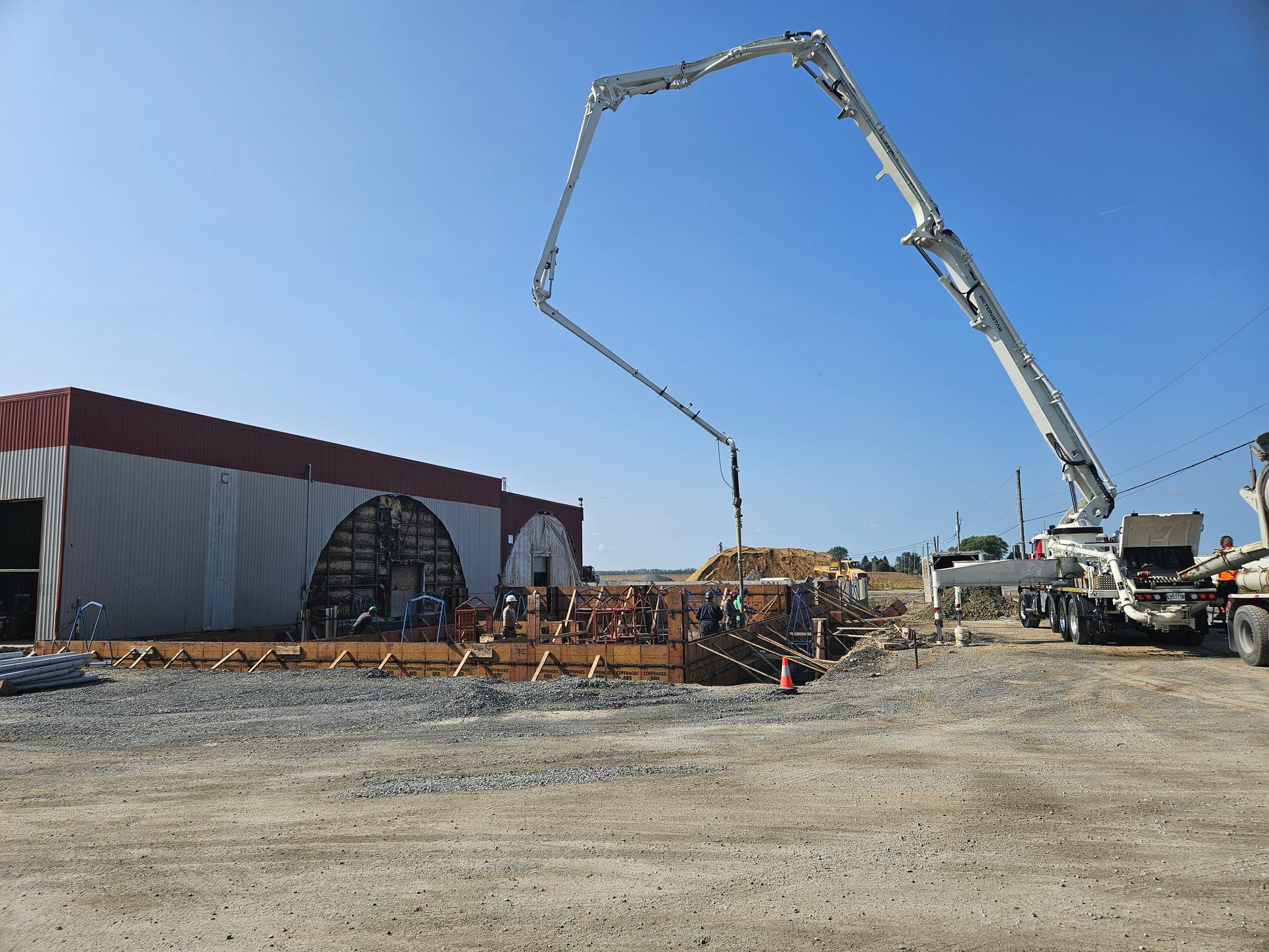 Un camion-pompe à béton étend son bras au-dessus d'une équipe de construction qui coule une fondation en béton près d'un grand bâtiment de stockage.