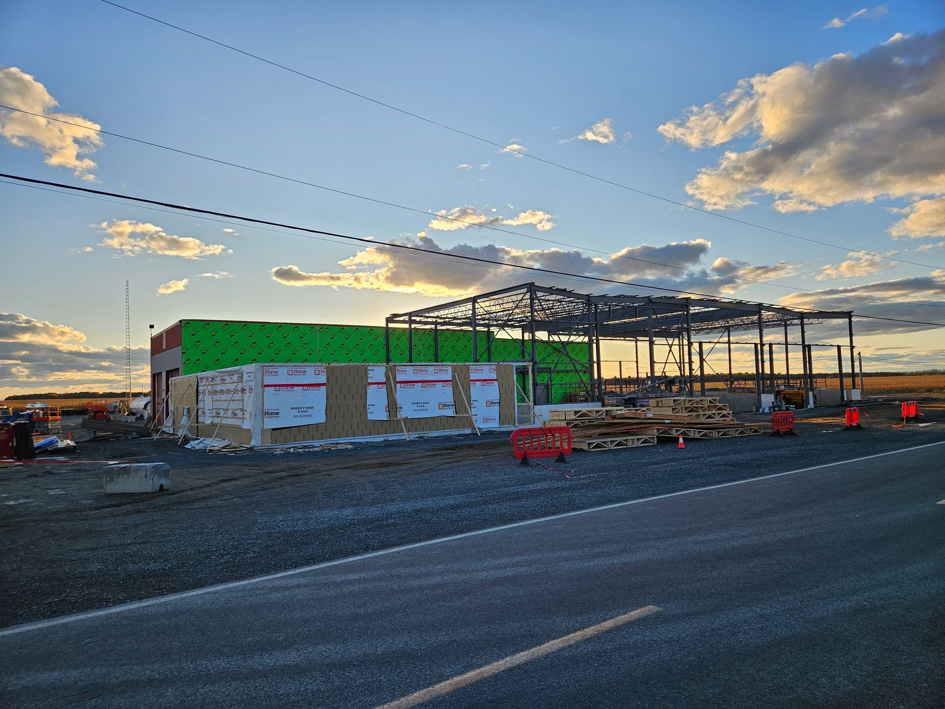 Chantier de construction au coucher du soleil, montrant une structure partiellement construite avec des panneaux extérieurs verts et une ossature en acier.