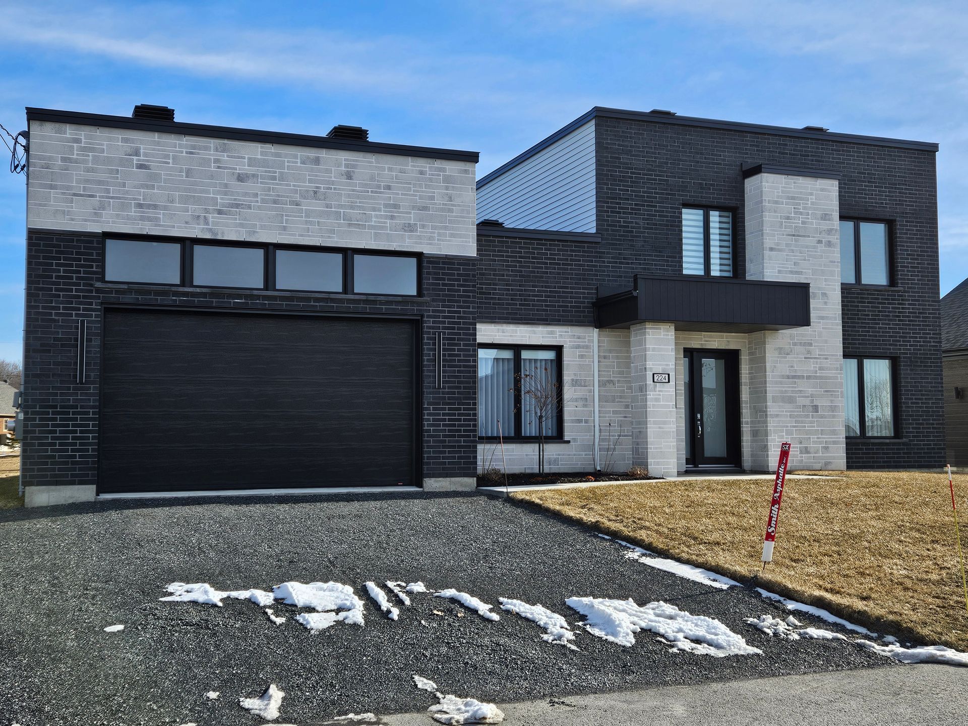 Une maison moderne à deux étages avec un mélange de revêtements en pierre noire et claire, une porte de garage foncée et une allée en gravier.