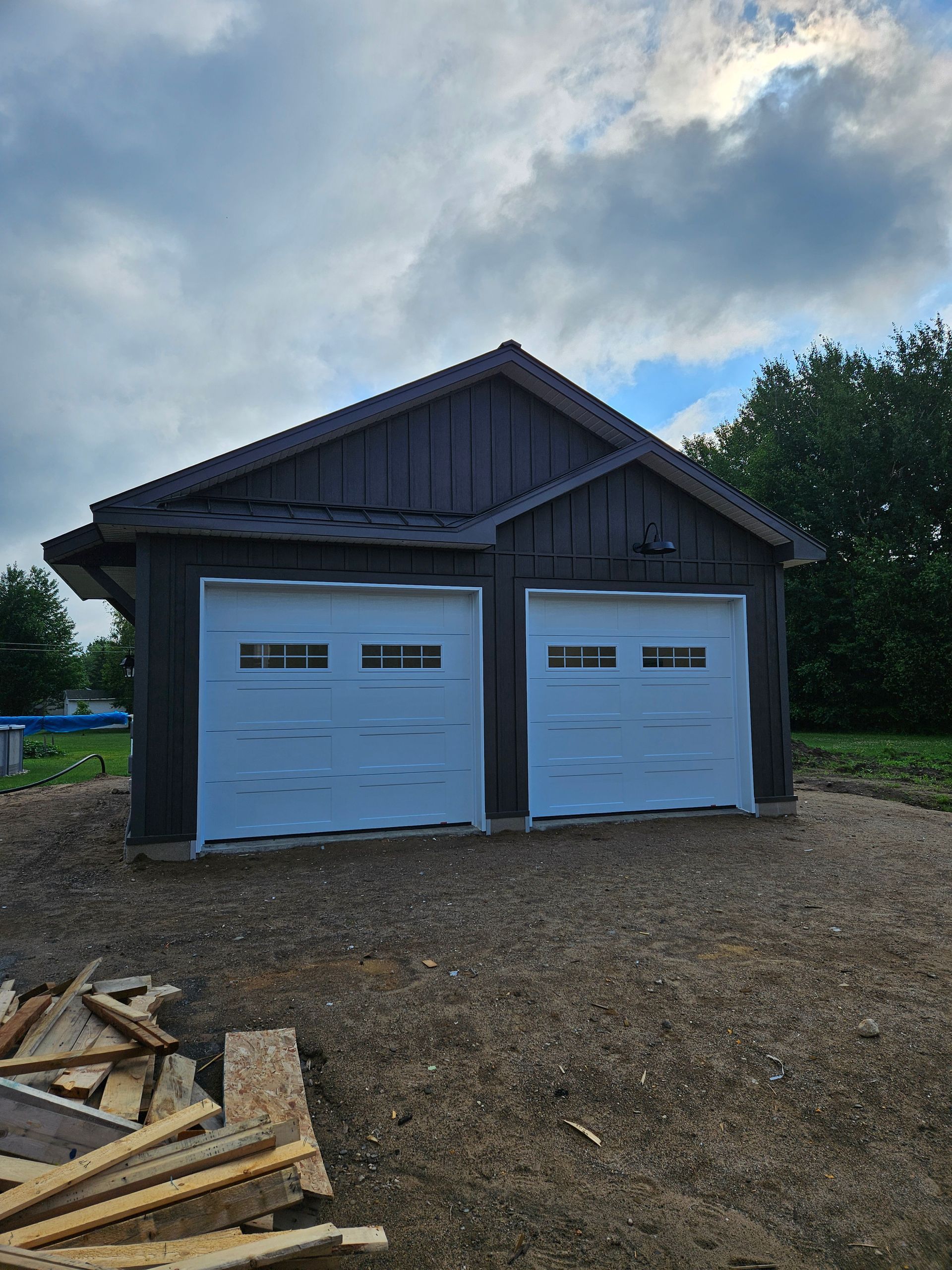 Un garage indépendant pour deux voitures, de couleur brun foncé et de construction récente, avec des portes blanches, sur un terrain gravillonné sous un ciel nuageux.