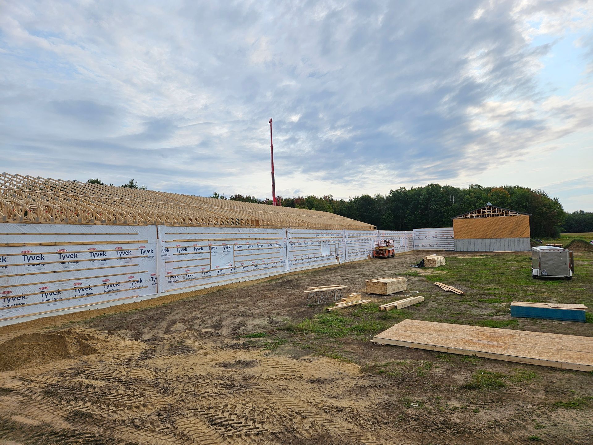 Un chantier de construction montrant une longue ossature murale recouverte d'un pare-vapeur blanc, avec des fermes de toit en bois et des matériaux de construction.