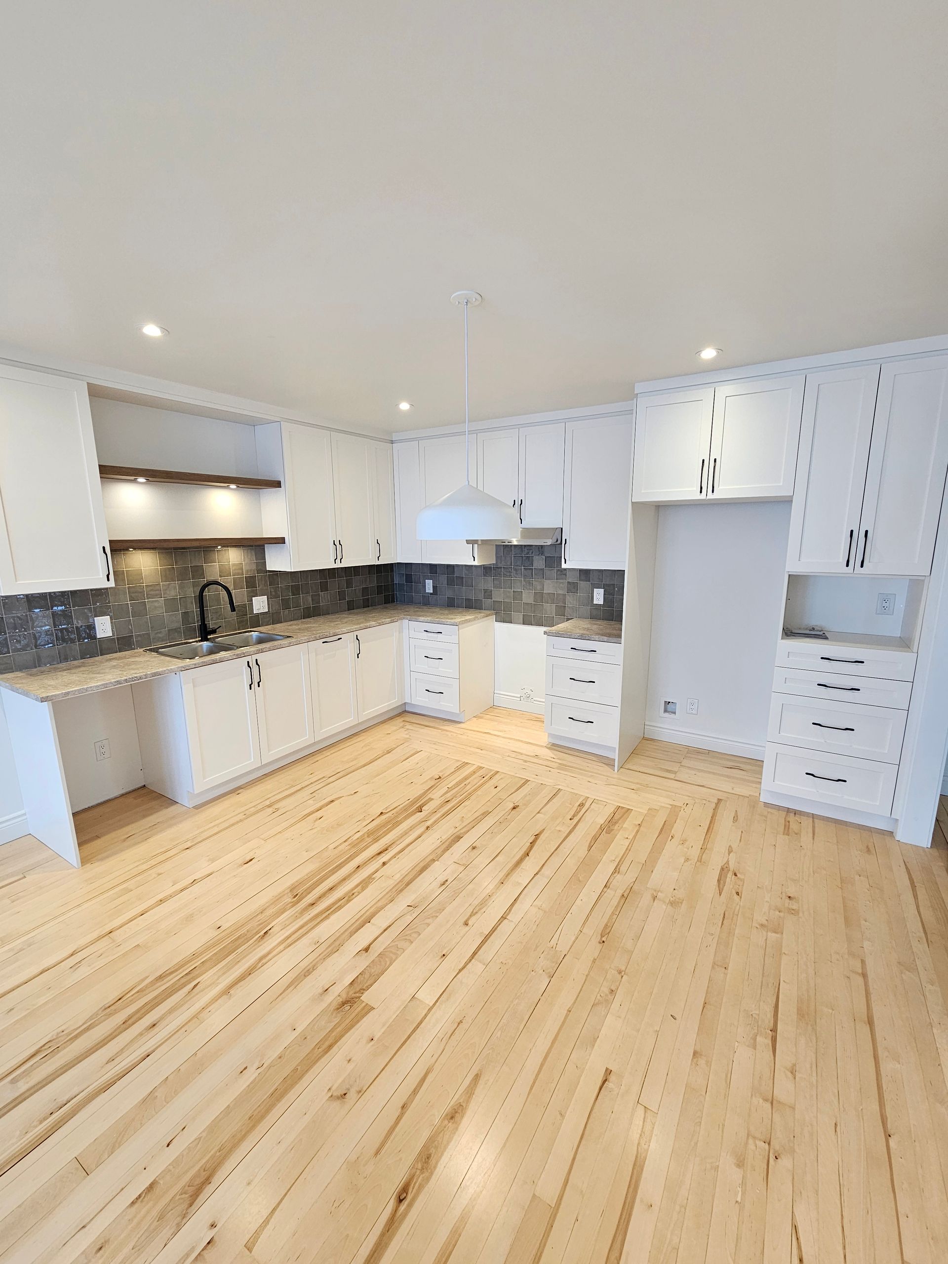 Une cuisine moderne et lumineuse avec des armoires blanches, une crédence en carrelage foncé, un parquet clair et un luminaire sur l'îlot central.