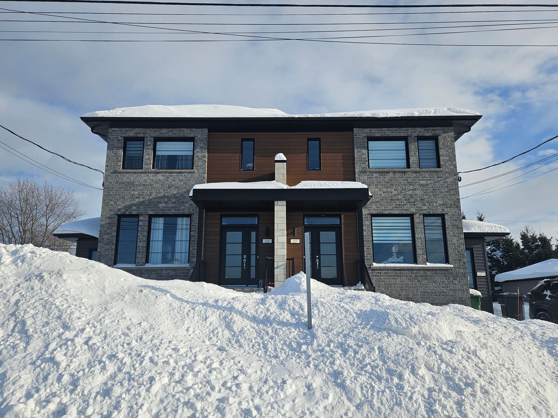 Un duplex moderne de deux étages, avec un revêtement en pierre grise et des accents de bois, entouré d'une épaisse couche de neige sous un ciel bleu.