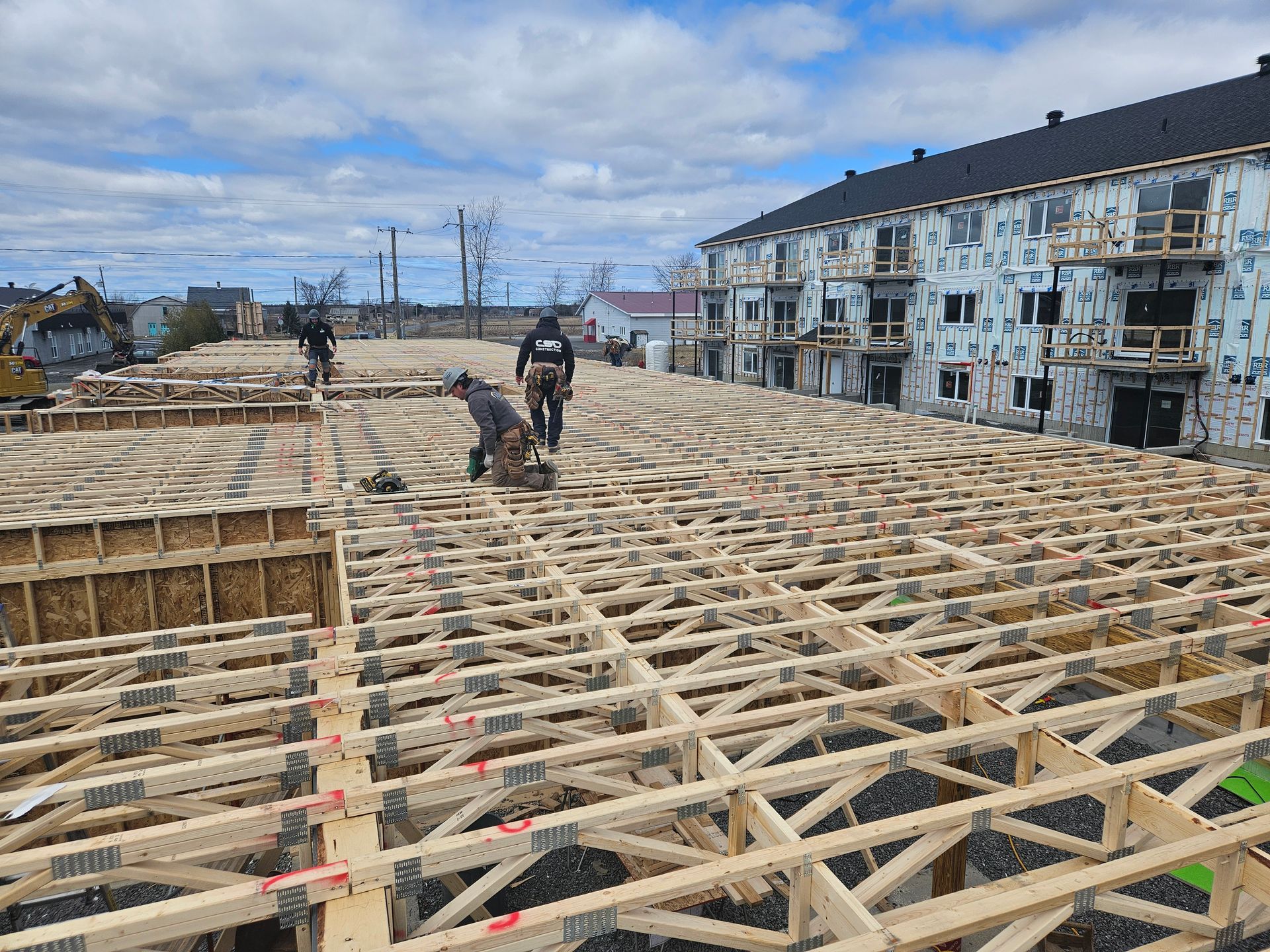 Des ouvriers du bâtiment installent des fermes de plancher en bois sur un grand chantier ouvert, jouxtant un immeuble d'appartements.
